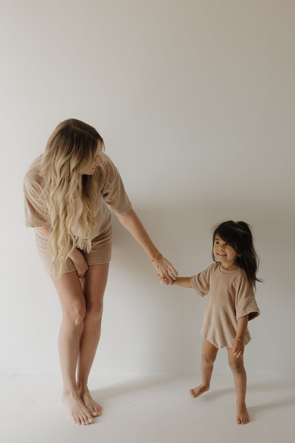 A woman with long blonde hair and a young girl smile at each other, both barefoot in beige outfits. The child wears the Tan Terry T-Shirt Romper by forever french baby, and they stand against a plain light-colored wall.