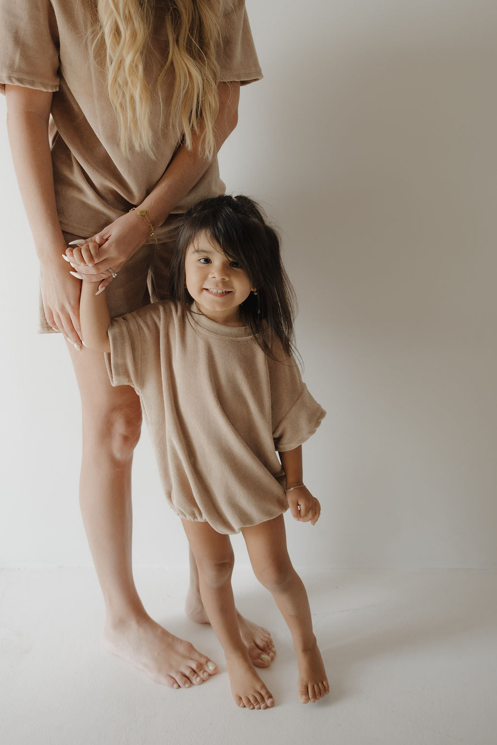 A young woman and a small child, both barefoot and wearing matching forever french baby Terry T-Shirt Rompers in Tan, stand against a plain white background. The woman gently holds the child’s hand as they share a sweet family moment.