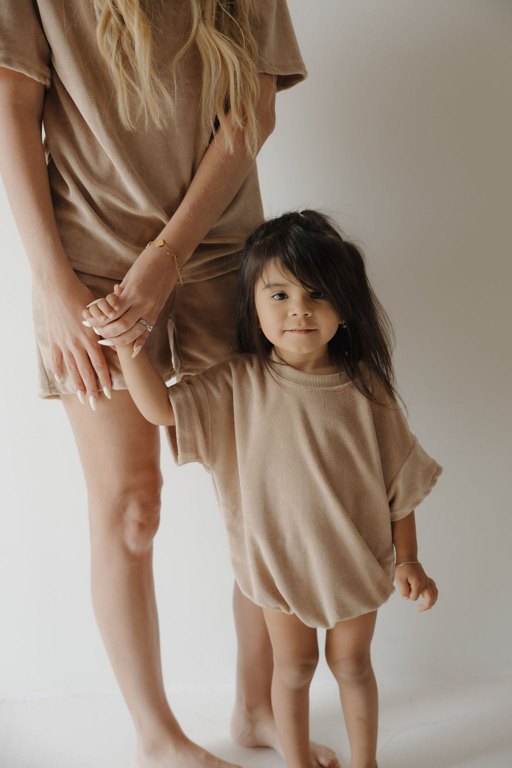 A woman and a young girl, barefoot against a white background, wear matching forever french baby Terry T-Shirt Rompers in Tan. The woman holds the girl's hand as they share a sweet family moment, with the girl smiling at the camera.