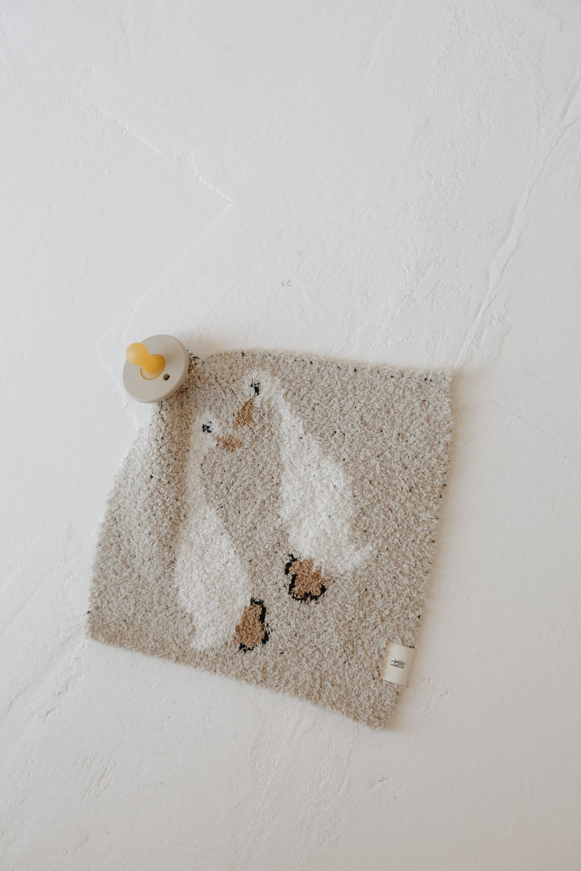 The Lovey | Silly Goose by forever french baby is a soft, square microfiber feather yarn washcloth featuring two white ducks. A yellow-and-gray pacifier rests on the upper left corner against a textured white background.