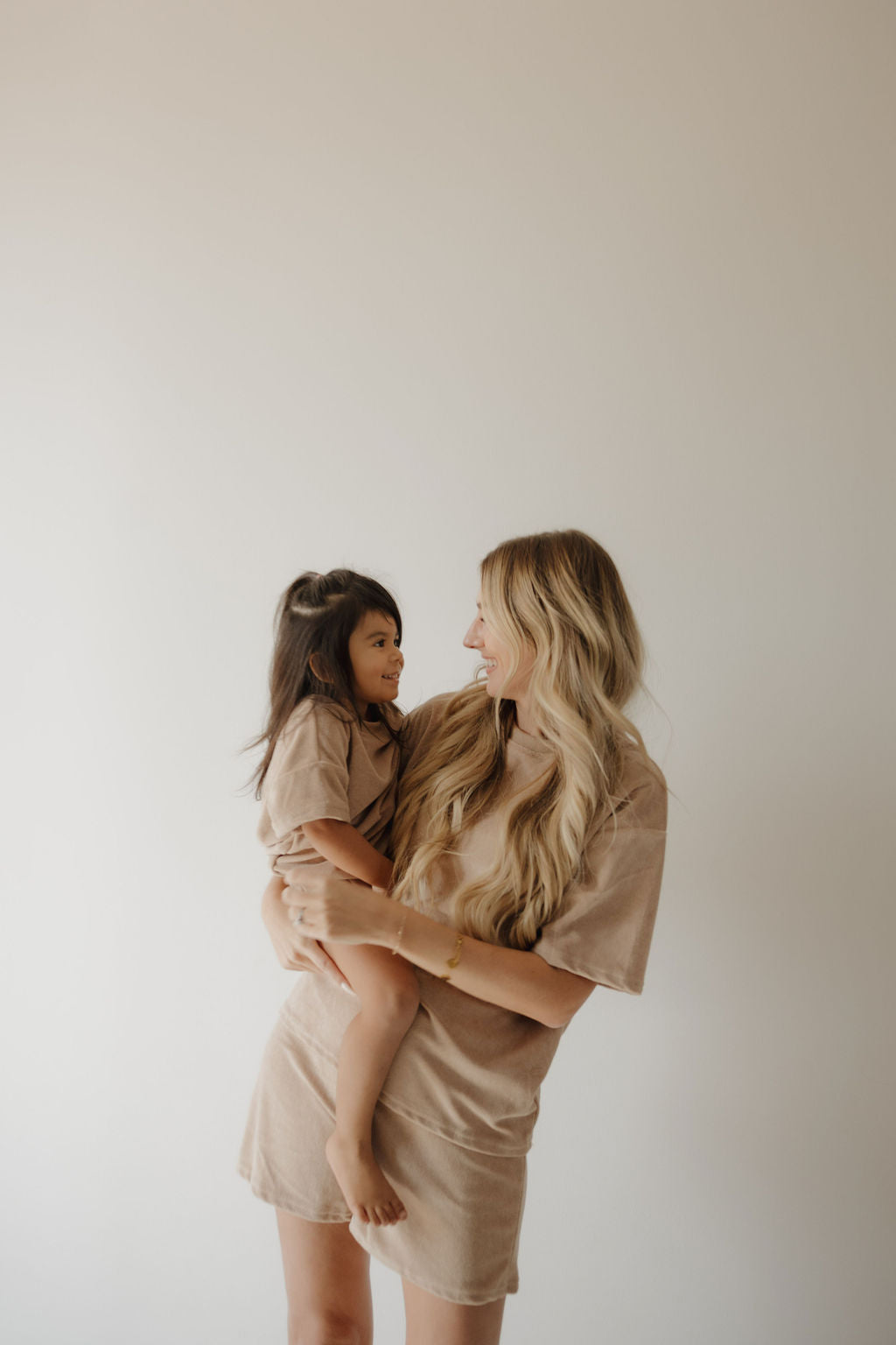 A woman holds a young girl as they smile at each other, both wearing forever french baby Terry T-Shirt Romper | Tan with snap closures. The matching outfits are perfect for newborns to 24 months, set against a plain, light background.