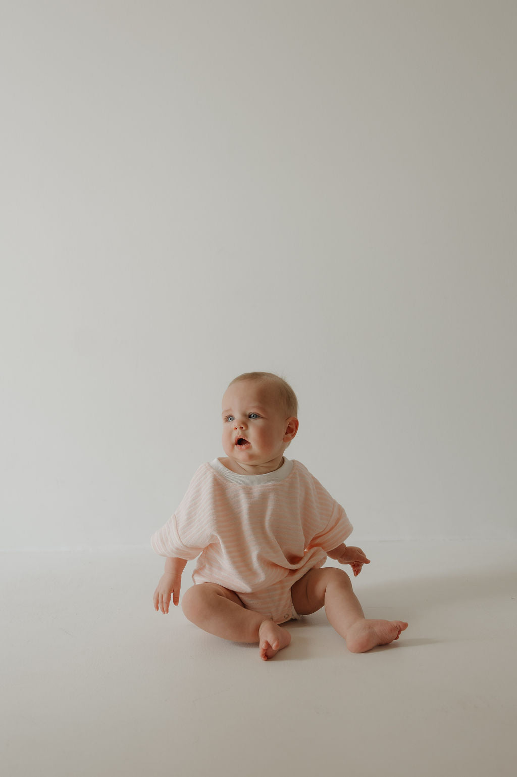A baby in the forever french baby Terry T-Shirt Romper | Cotton Candy with pink stripes and snap closure sits on a plain white background, looking to the side—perfect for capturing sweet family moments.
