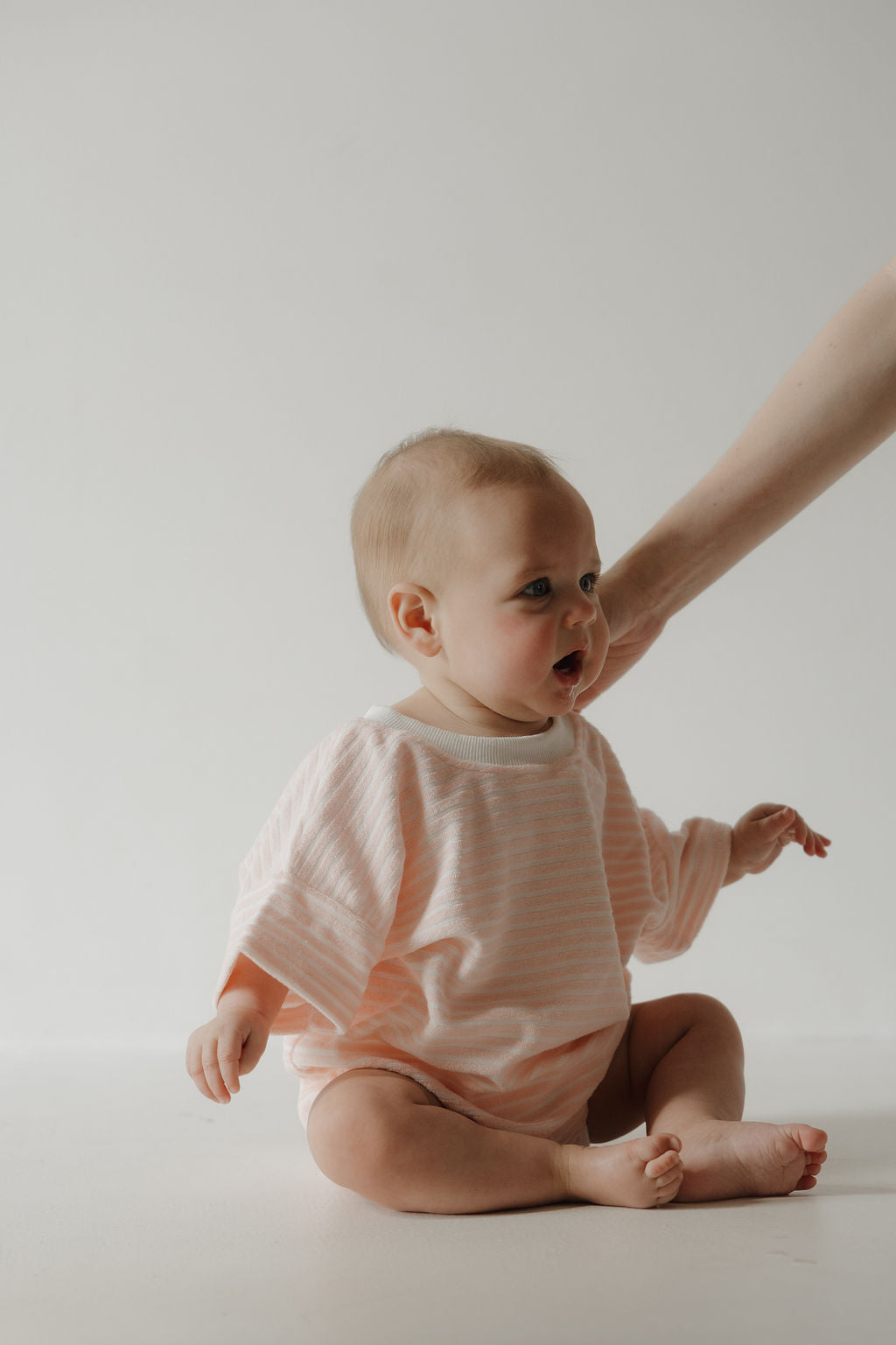A baby in the forever french baby Terry T-Shirt Romper | Cotton Candy sits on the floor against a white background, with an adult’s arm gently supporting them for easy, comfortable diaper changes.