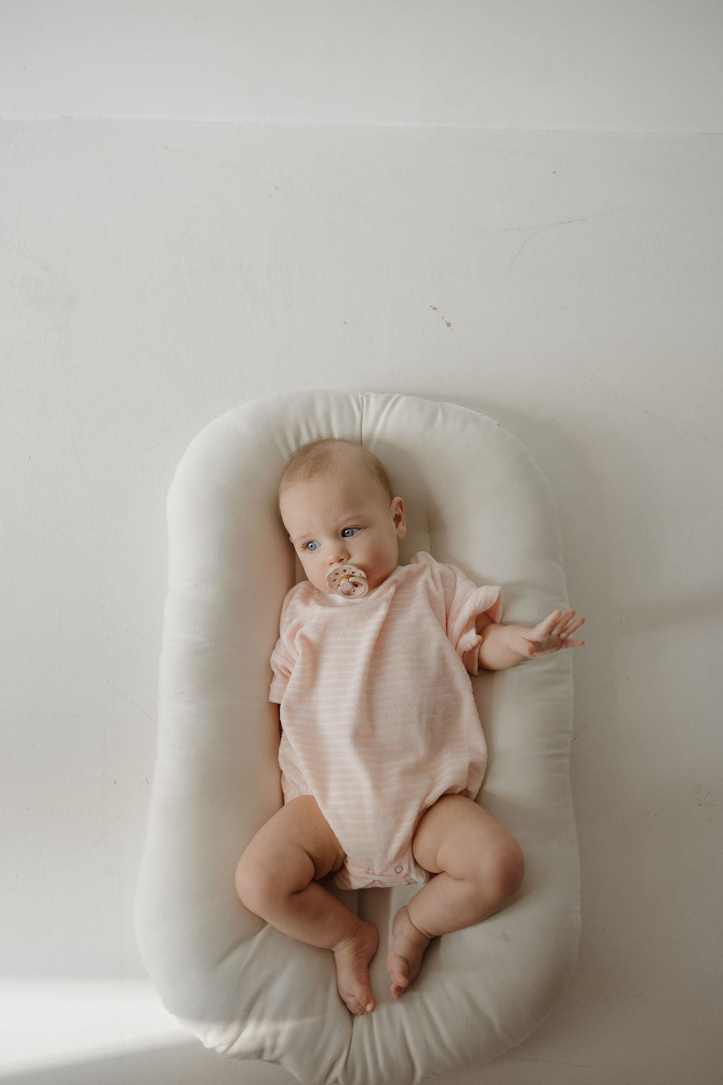 A baby with a pacifier lies on a white cushioned pad, wearing the forever french baby Terry T-Shirt Romper in Cotton Candy, perfect for easy diaper changes. The background is plain white.