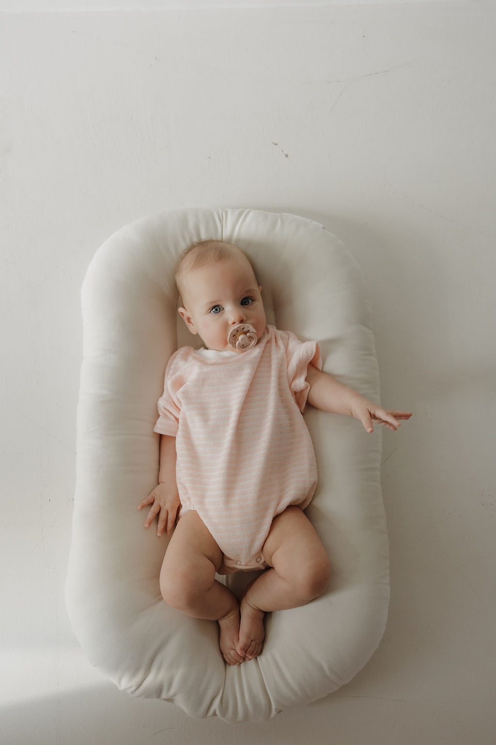 A baby with a pacifier lies on a soft white cushion, wearing the forever french baby Terry T-Shirt Romper | Cotton Candy, light pink striped with snap closure—perfect for cherishing sweet family moments.