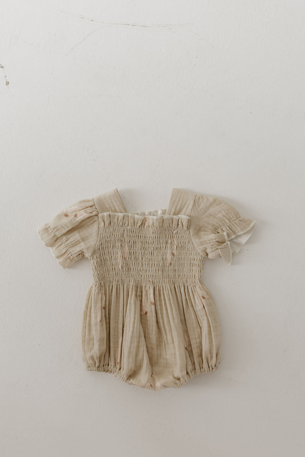 The forever french baby Ruffle Muslin Romper | Silly Goose is a beige romper with short puffed sleeves, ruffles, smocked bodice, elastic leg openings, and snap closure. Shown on a plain light background.