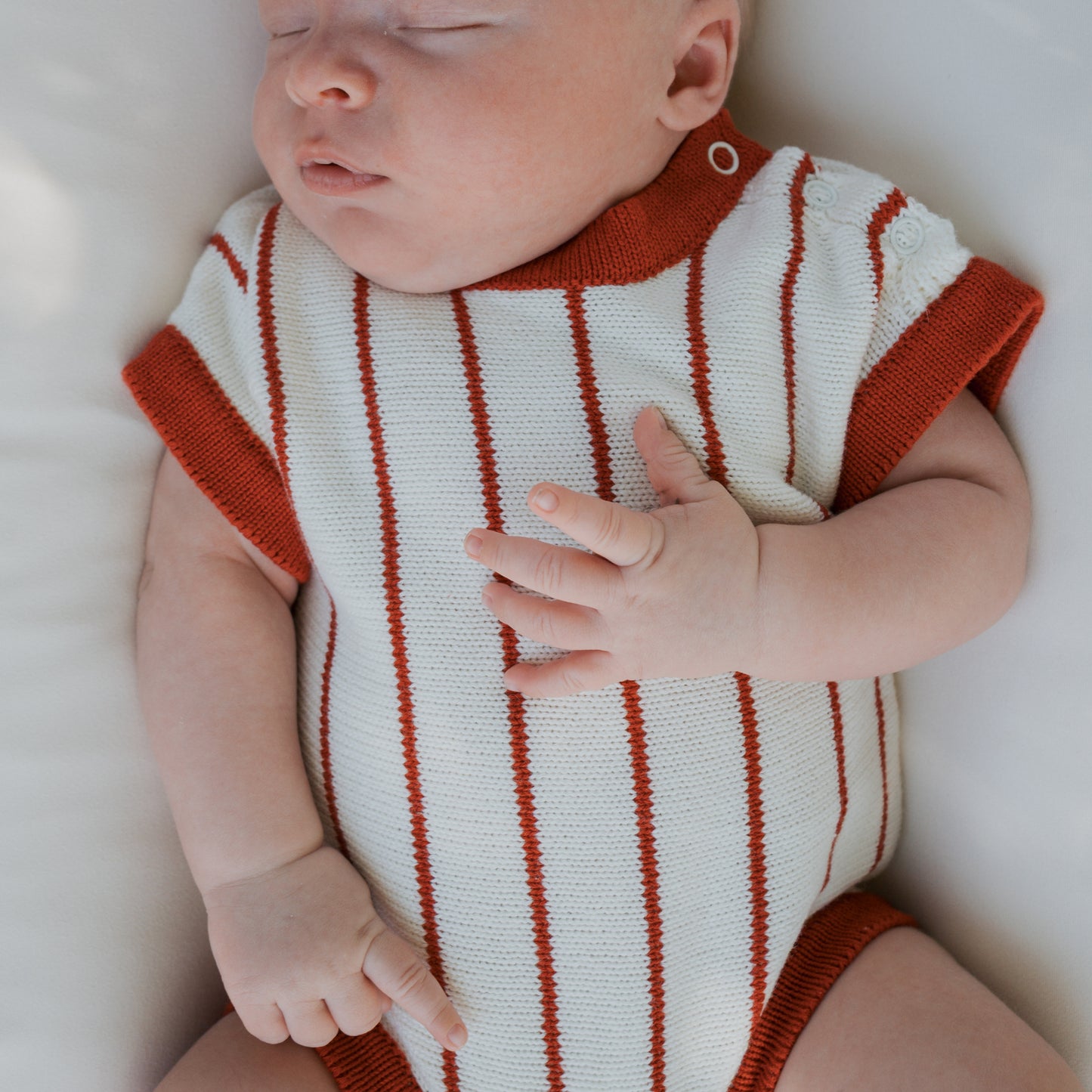 A newborn sleeps peacefully in the forever french baby Knit Romper | Picnic Pinstripe, featuring soft red stripes and trim, on a cozy white surface.