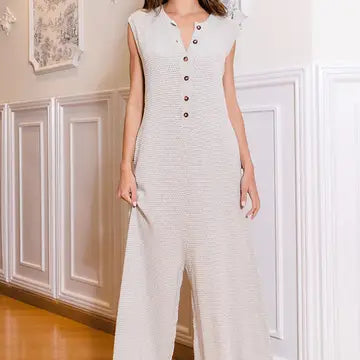 A woman stands indoors wearing the Wide Leg Button-Up Loose Fit Solid Sweater Jumpsuit by Bucketlist. The room features white paneled walls and framed artwork, creating a cozy backdrop that complements her loose-fit ensemble.