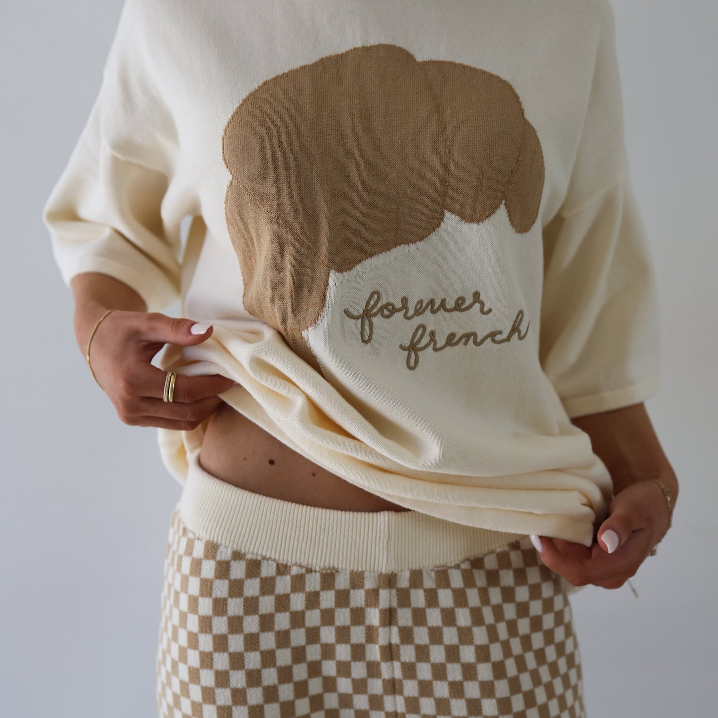 A person models the Women's Knit Pant Set | Croissants from forever french baby, featuring an oversized cotton sweater with a brown croissant graphic and "forever french," paired with beige and white checkered knit shorts.