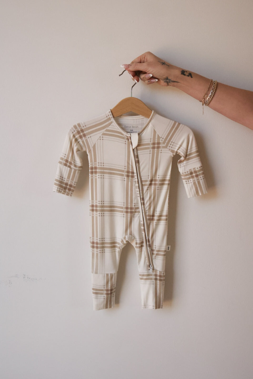 A hand holds forever french baby's Bamboo Zip Pajamas | Ellis Plaid, a beige and white plaid long-sleeve baby romper made from breathable fabric, against a plain light-colored wall.