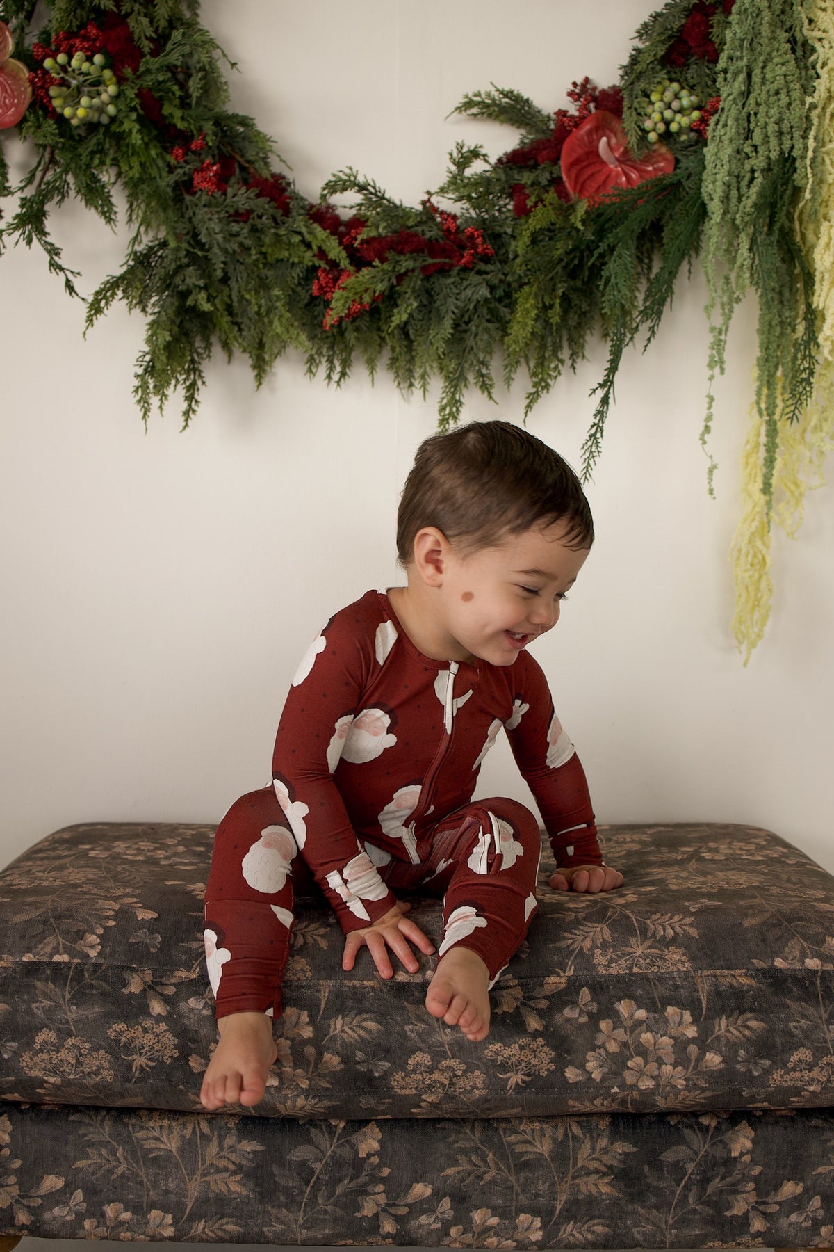 A toddler wearing dark red Bamboo Zip Pajamas | Vintage Santa, sitting on a floral ottoman.