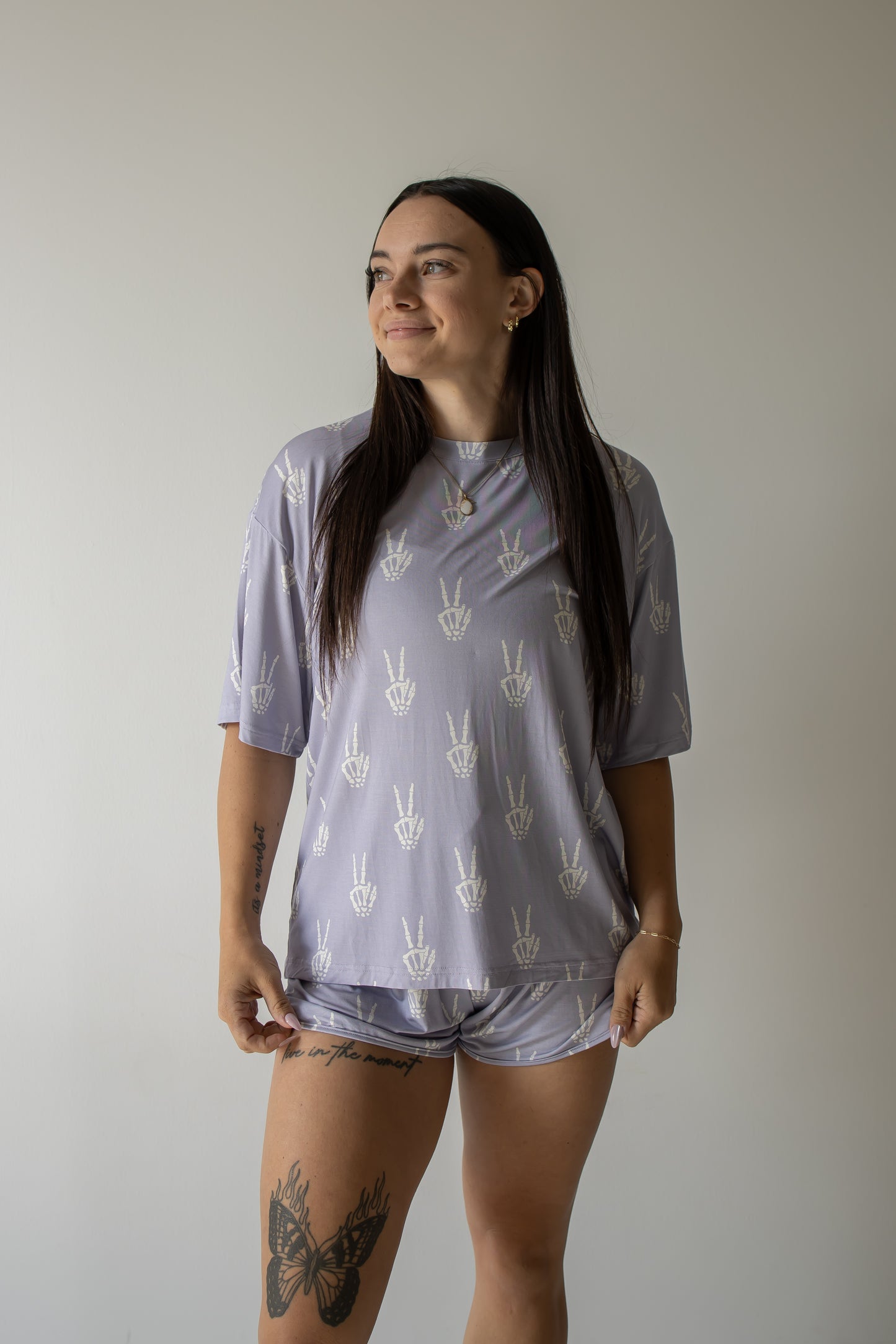 A woman with long dark hair, tattoos on her thigh and forearm, stands smiling and looking to the side against a plain light background. She is wearing the forever french baby Women’s Bamboo Short Set | Enchanted Skelly— a matching lavender pajama set with white hand symbols—highlighting the soft and hypo-allergenic comfort of bamboo clothing.