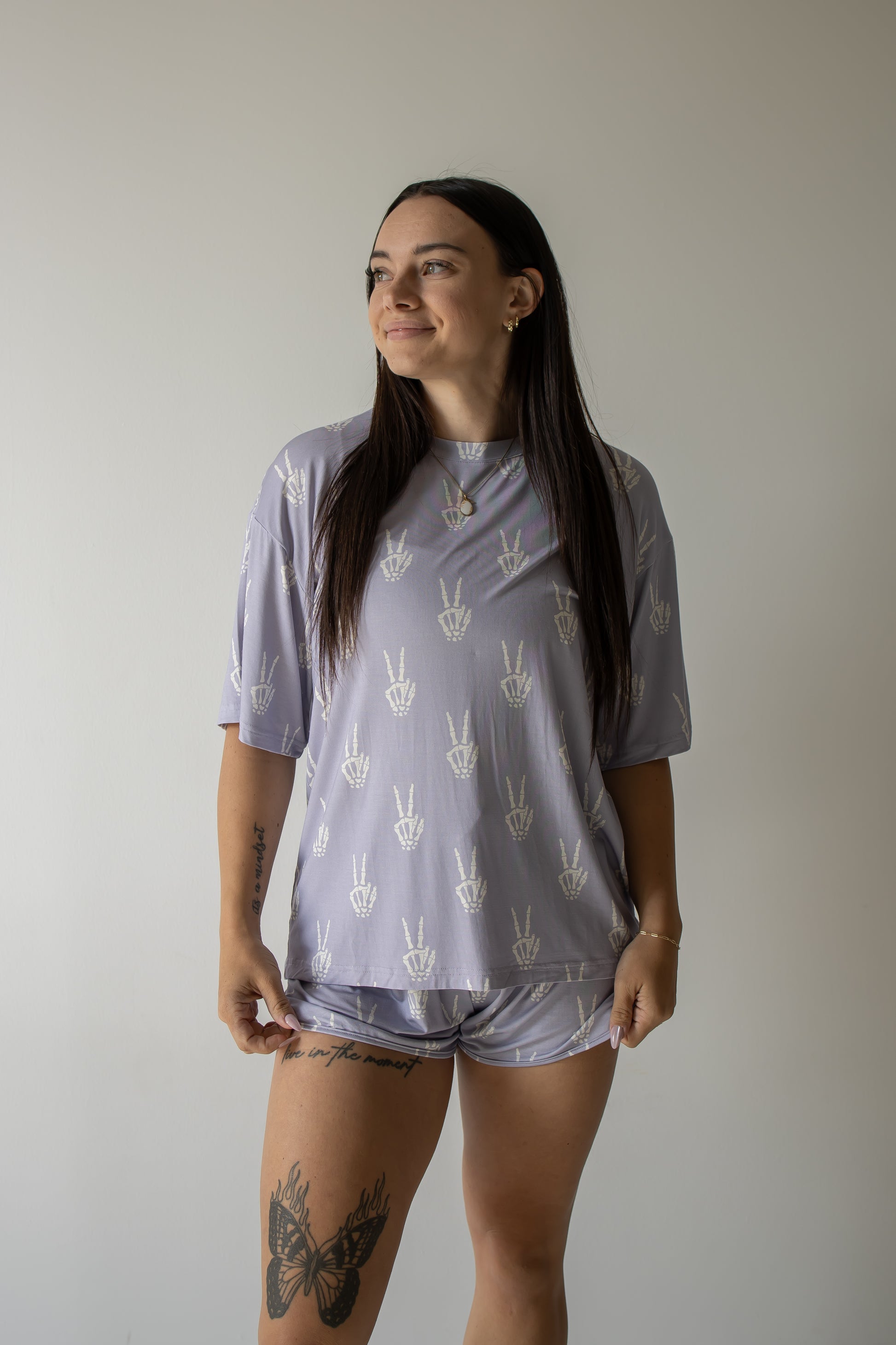 A woman with long dark hair, tattoos on her thigh and forearm, stands smiling and looking to the side against a plain light background. She is wearing the forever french baby Women’s Bamboo Short Set | Enchanted Skelly— a matching lavender pajama set with white hand symbols—highlighting the soft and hypo-allergenic comfort of bamboo clothing.