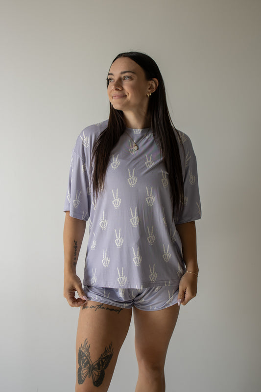 A woman with long dark hair, tattoos on her thigh and forearm, stands smiling and looking to the side against a plain light background. She is wearing the forever french baby Women’s Bamboo Short Set | Enchanted Skelly— a matching lavender pajama set with white hand symbols—highlighting the soft and hypo-allergenic comfort of bamboo clothing.