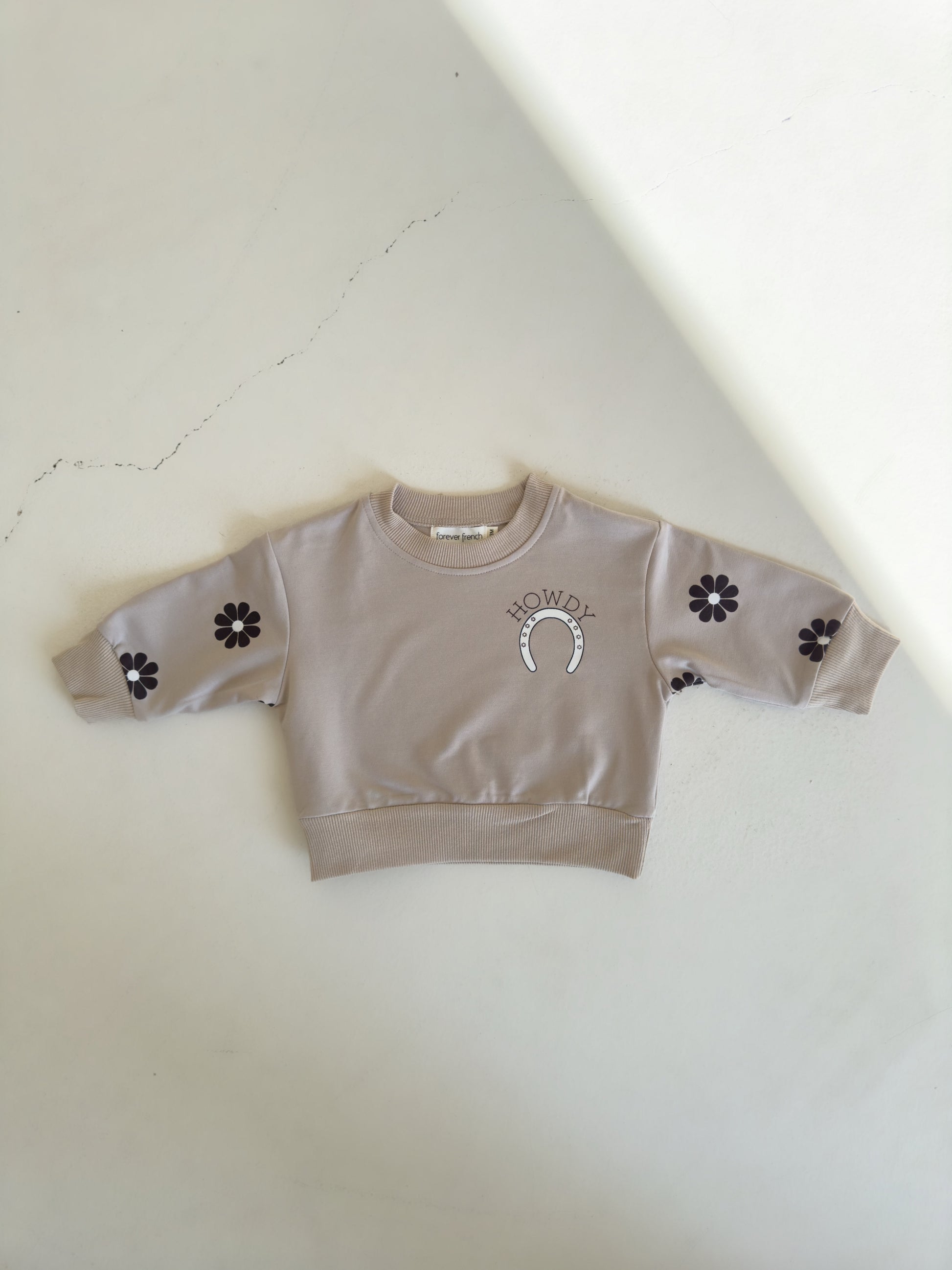 Part of the Howdy Collection by forever french baby, this beige Child Sweatshirt features black flower designs on the sleeves and "HOWDY" with a charming horseshoe graphic on the front, set against a cracked light-colored surface to create a matching moment in your wardrobe.