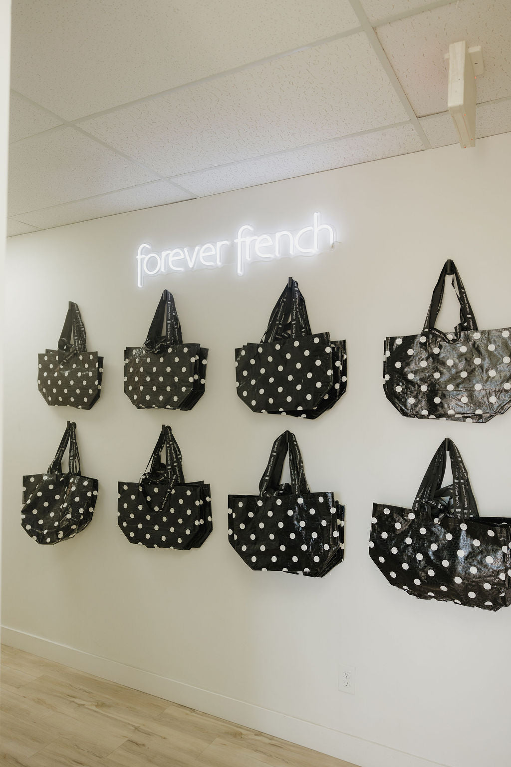 Eight ff Warehouse Sale Tote Bags in black and white gingham from forever french baby are displayed on a white wall beneath a glowing “forever french” sign. The room features a light wood floor and a white ceiling.