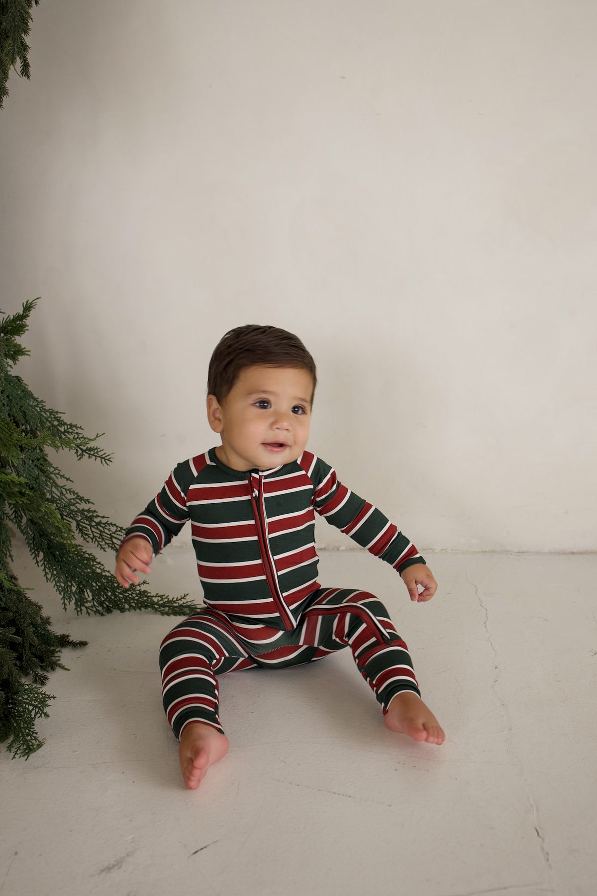 A baby sits on the floor wearing Bamboo Zip Pajamas | Holiday Stripe, featuring red, white, and green horizontal stripes and a center zipper.
