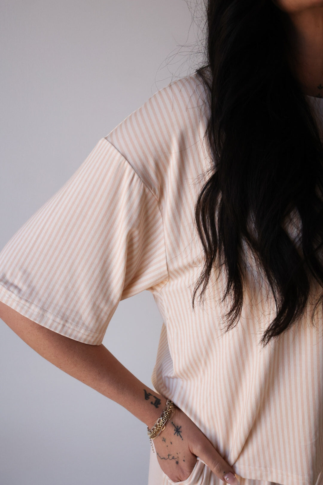 A woman with long dark hair wears the forever french baby Women's Boxy Tee Lounge Set in Sadie Stripe—a short-sleeved, light, vertically-striped set—standing with one hand on her hip, displaying bracelets and finger tattoos.