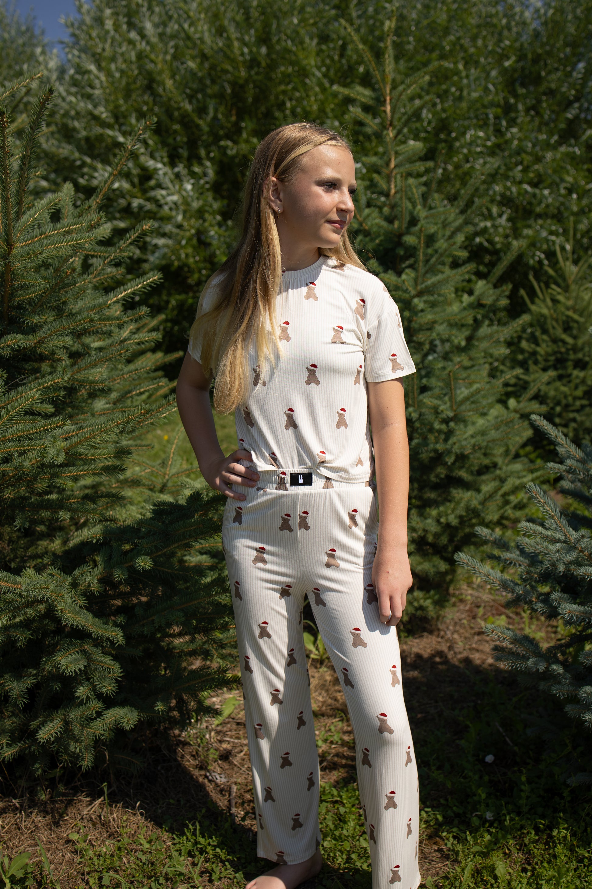 A girl stands outdoors among green pine trees, wearing the forever french baby Child Boxy Tee Lounge Set in the Santa Teddy design—a matching cream-colored pajama set with small brown figure prints. Crafted from soft, hypo-allergenic bamboo fabric, her outfit complements her long blonde hair as she rests one hand on her hip, gazing off to the side in the sunlight.