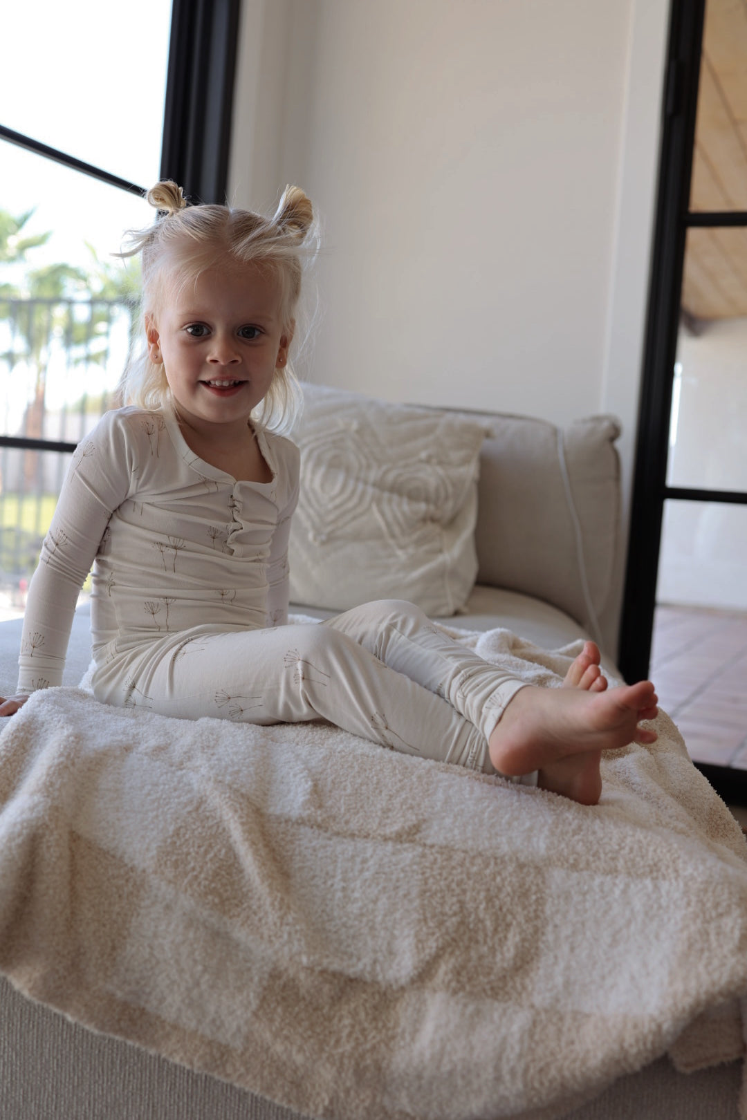 A blonde-haired young child with two buns sits barefoot on a light couch, smiling at the camera. She wears forever french baby Bamboo Two Piece Pajamas | Dandelion. Large windows and greenery are visible in the background.