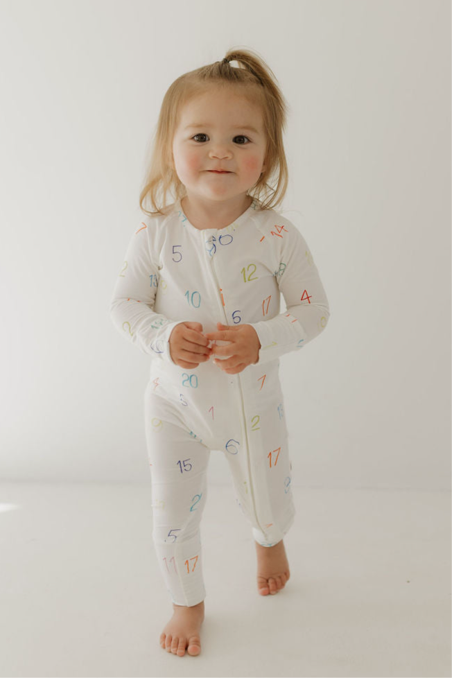 A toddler with light brown hair in a ponytail wears forever french baby Bamboo Zip Pajamas | Autism Awareness | Charli Counts—hypo-allergenic white pajamas featuring colorful numbers—standing and smiling against a plain white background.