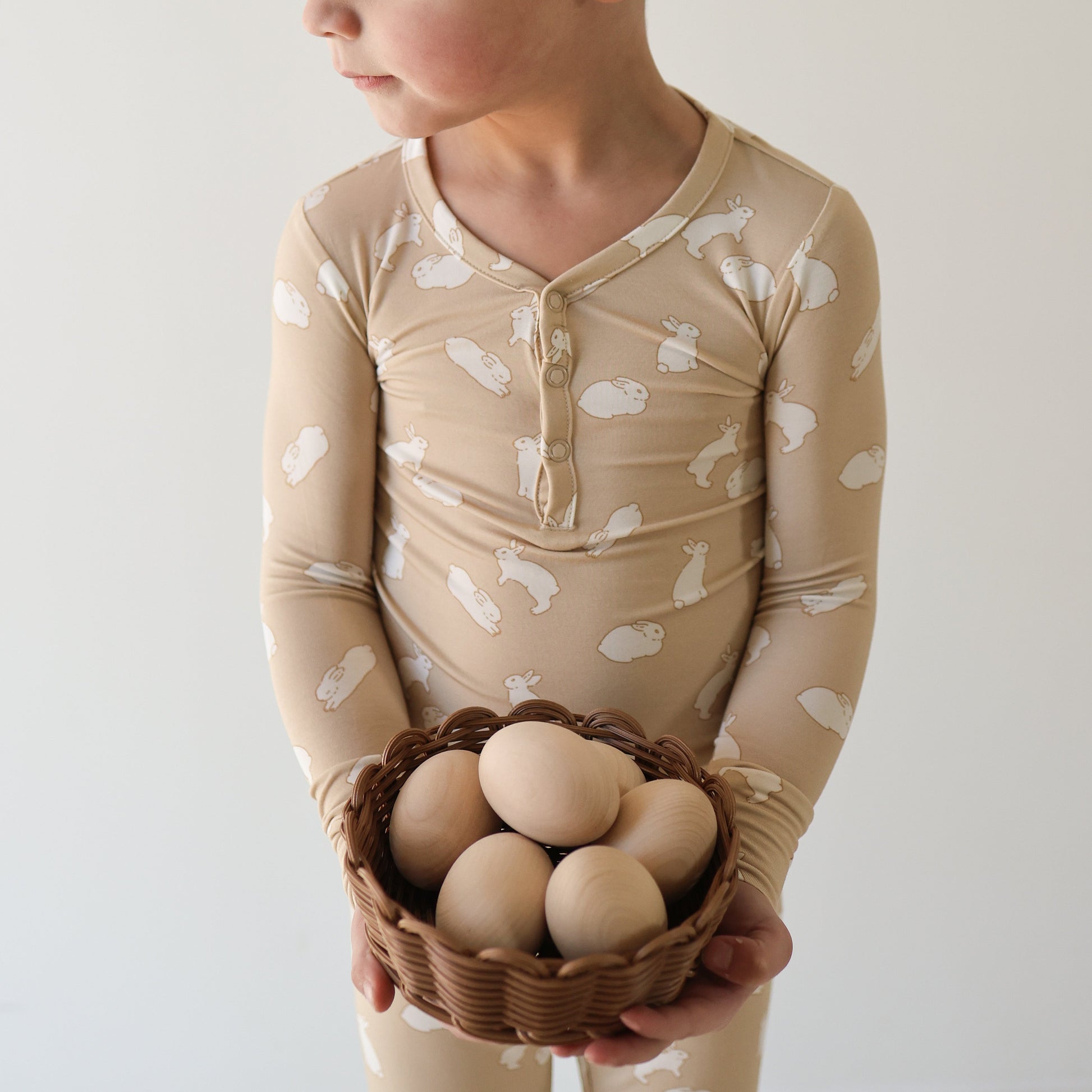 A child wears forever french baby's Bamboo Two Piece Pajamas | Bunnies—beige with white bunny prints—while holding a woven basket of wooden eggs against a light background.