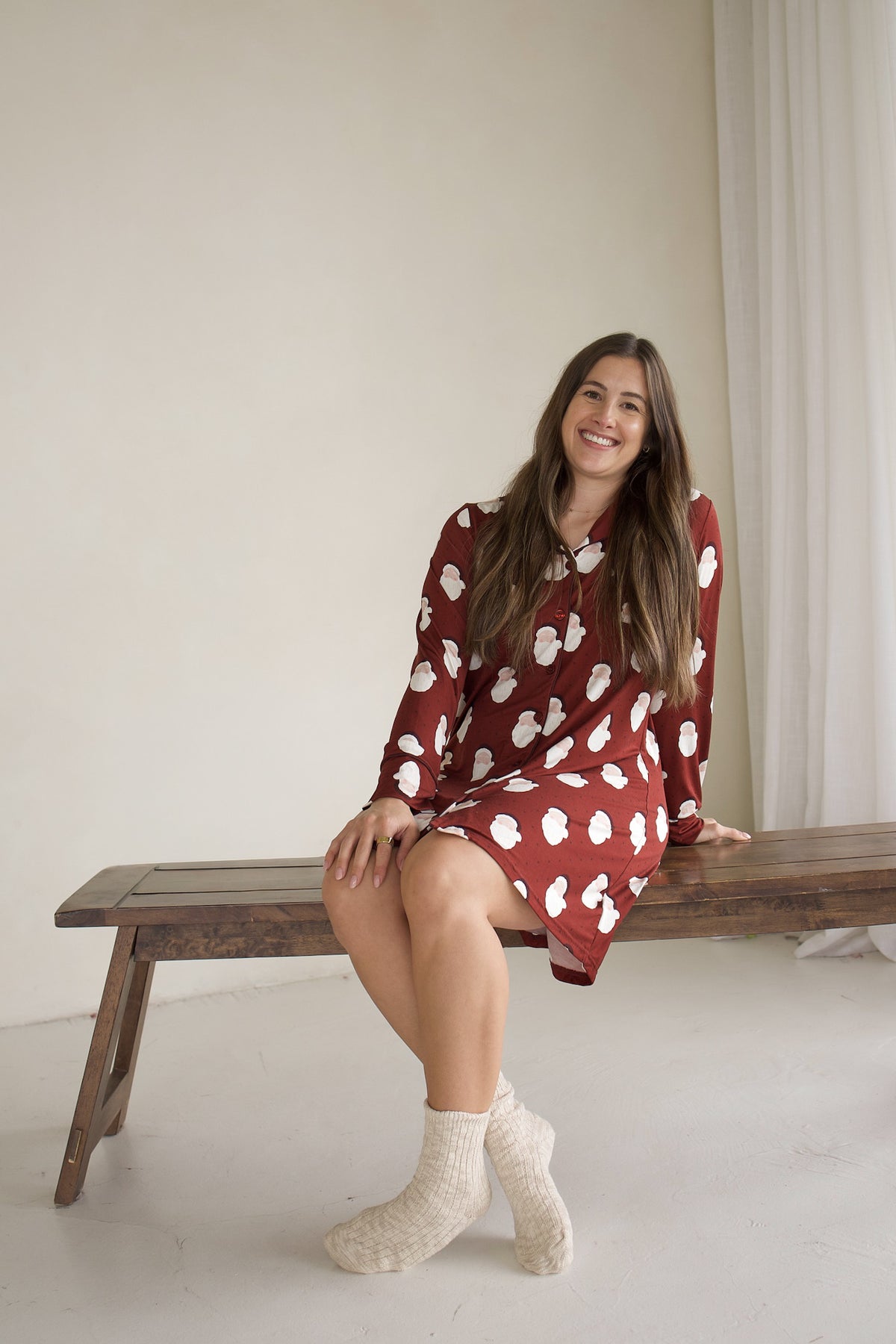 A smiling woman sits on a wooden bench wearing a maroon Women’s Bamboo Sleeping Dress | Vintage Santa with a white cloud print and cream-colored socks.