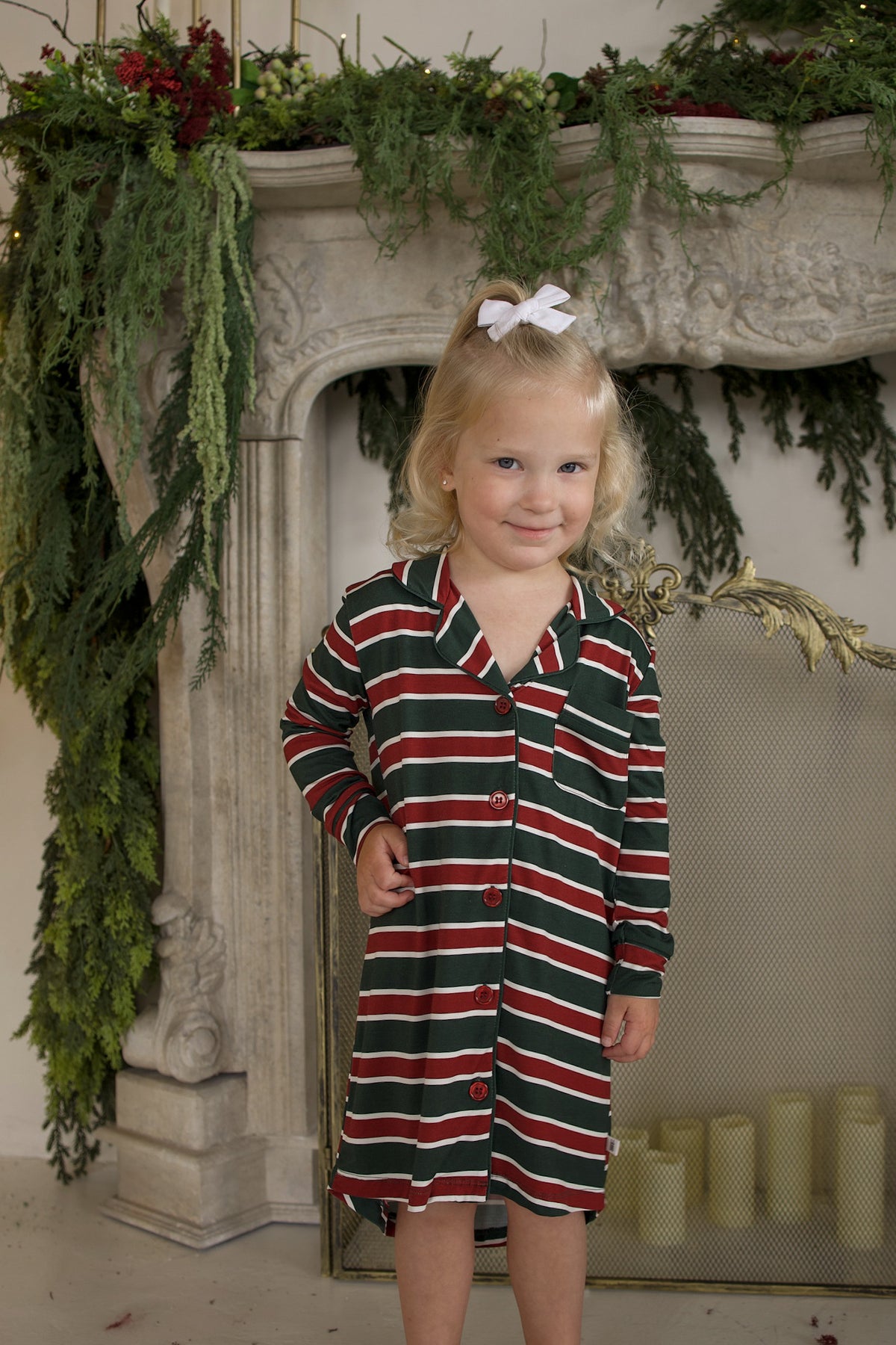 A young girl in the Bamboo Sleeping Dress | Holiday Stripe by forever french baby stands before a decorated fireplace.