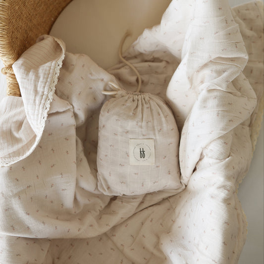 The Muslin Crib Sheet | Lucy by forever french baby, in a soft, light hue with a dotted pattern, is displayed beside a wicker basket. A matching “bb” drawstring bag rests on the sheet.