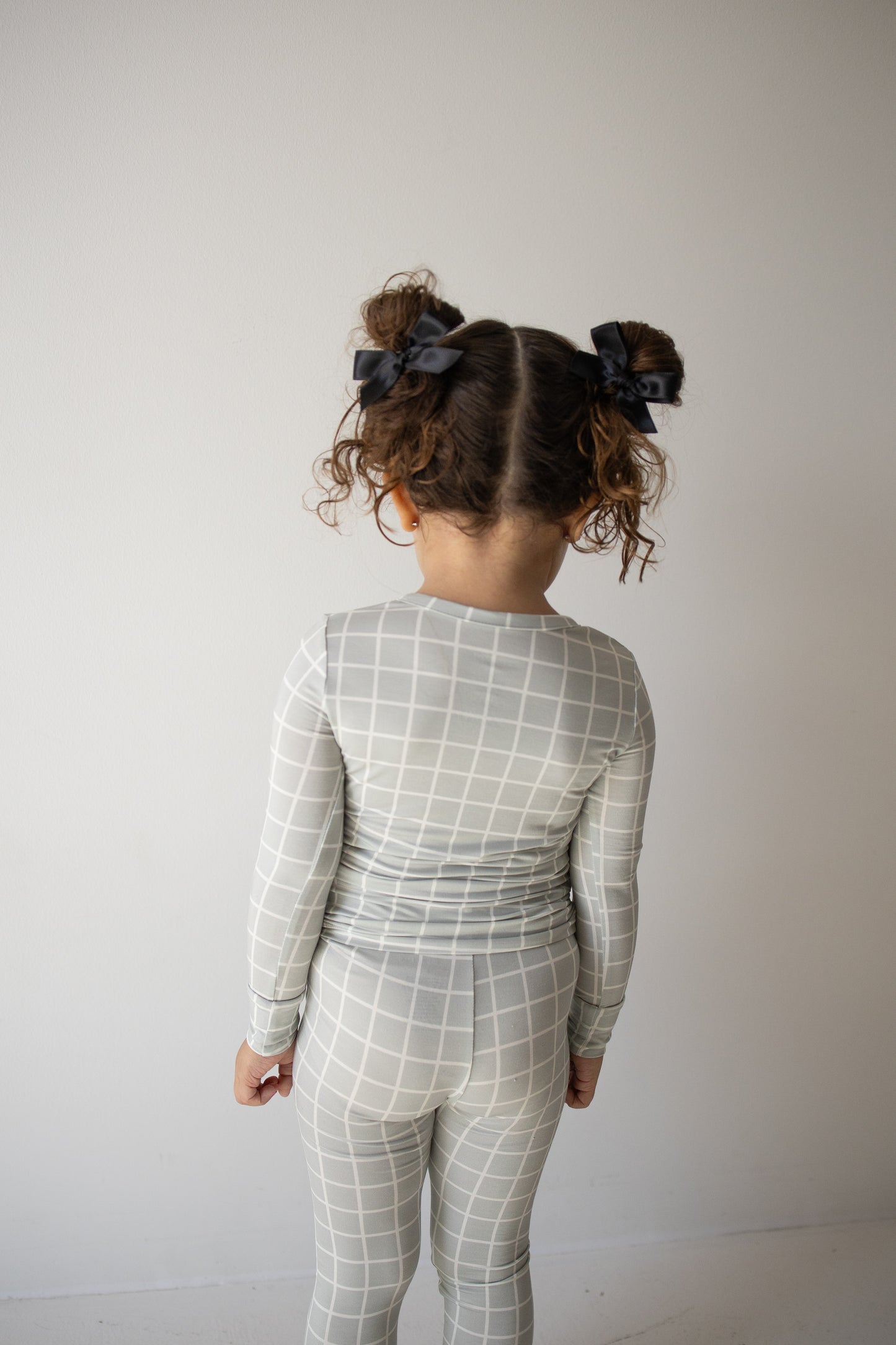 A young child with curly hair in pigtails, wearing the forever french baby Bamboo Two Piece Pajamas in the Foggy Grid pattern—featuring a matching gray and white grid-patterned, long-sleeve top and pants crafted from breathable bamboo fabric—stands facing away from the camera against a plain white background.