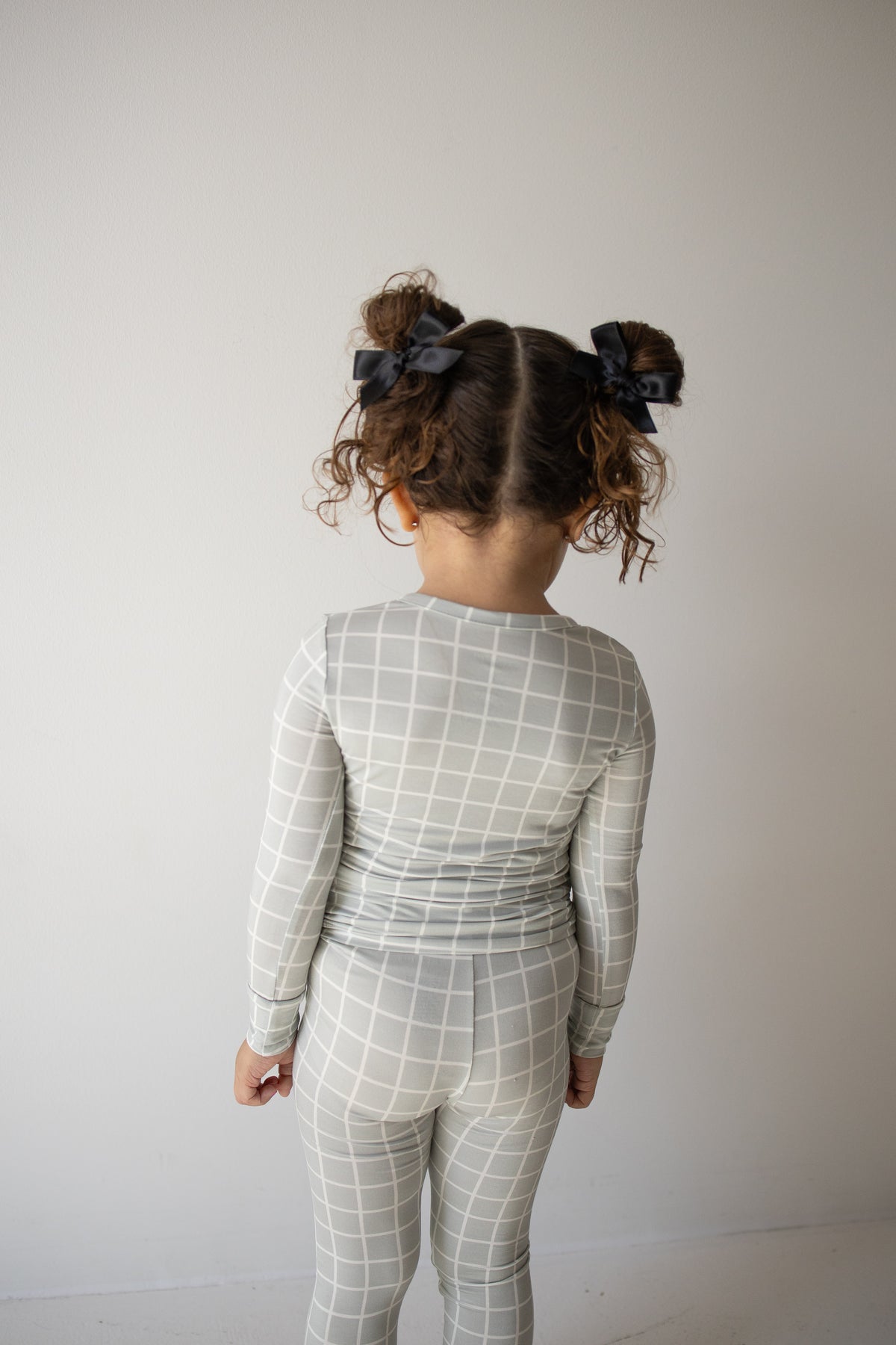 A young child with curly hair in pigtails, wearing the forever french baby Bamboo Two Piece Pajamas in the Foggy Grid pattern—featuring a matching gray and white grid-patterned, long-sleeve top and pants crafted from breathable bamboo fabric—stands facing away from the camera against a plain white background.