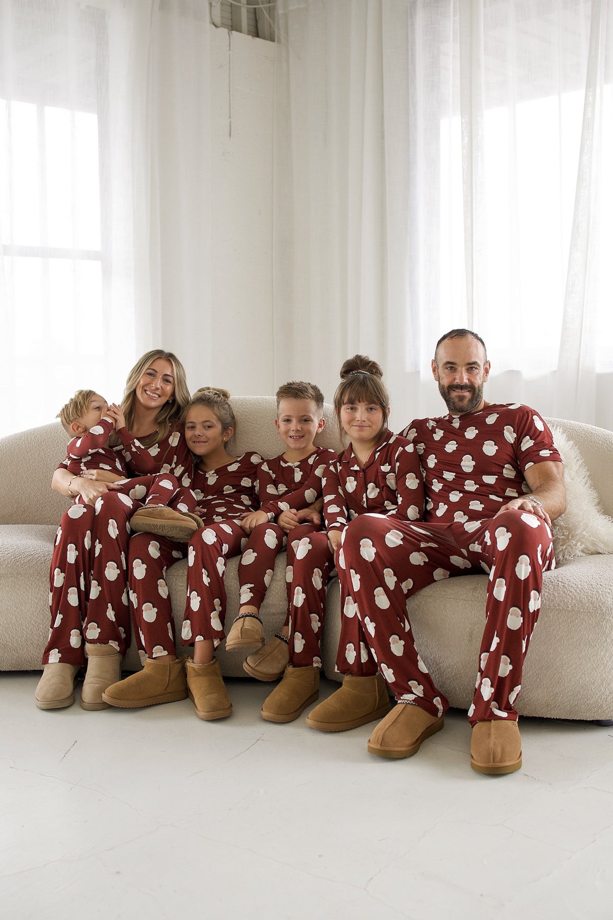 A family of six sits on a sofa wearing matching maroon Men’s Short Sleeve Pajamas | Vintage Santa bamboo sets with a white Santa Claus print and tan slip-on boots.