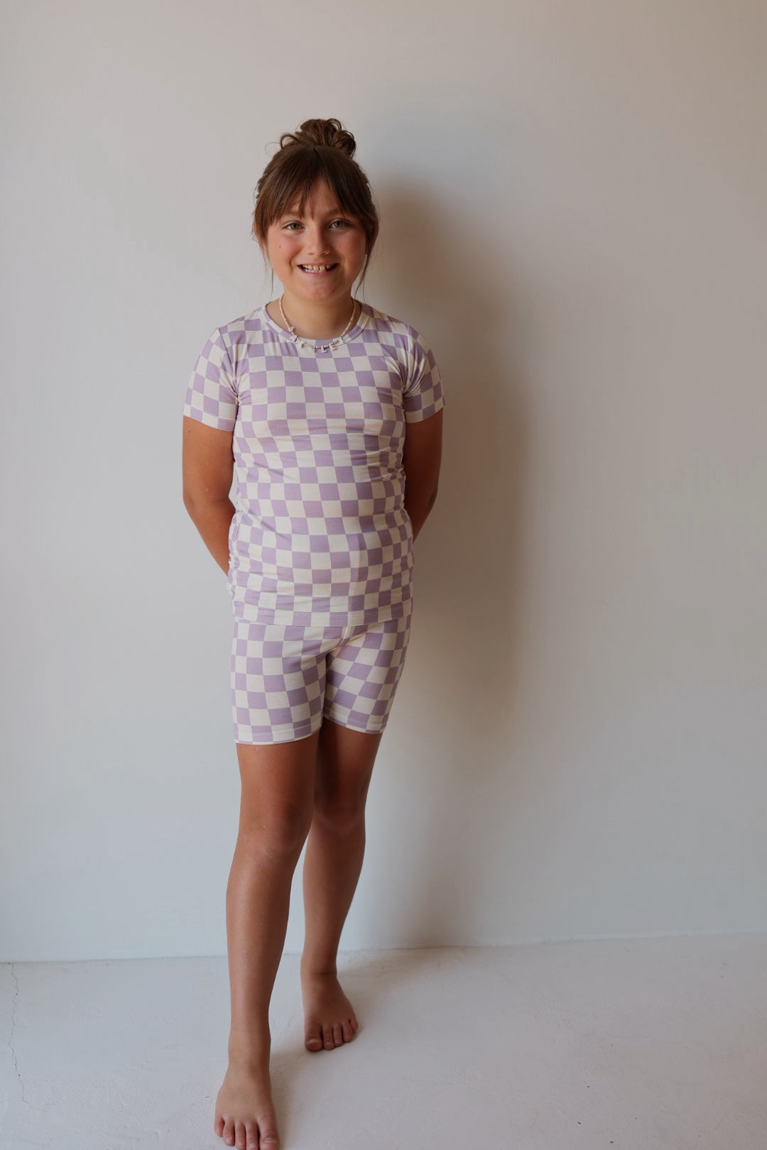 A young girl stands barefoot, smiling with her hands behind her back. She wears the forever french baby Bamboo Short Set | Grape Soda—comfortable, breathable sleepwear in a purple and white checkerboard print, perfect for relaxing or bedtime.