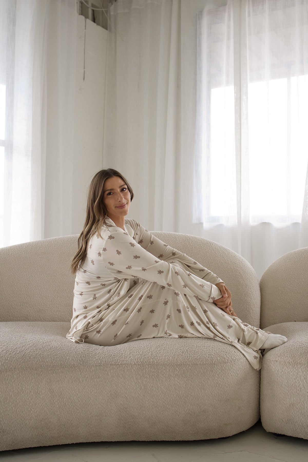 A woman sits on a white sofa wearing a Women’s Bamboo Button Up Lounge Set | Gingersnap featuring a long-sleeved button-up shirt and wide-leg lounge pants with a small gingerbread man pattern.
