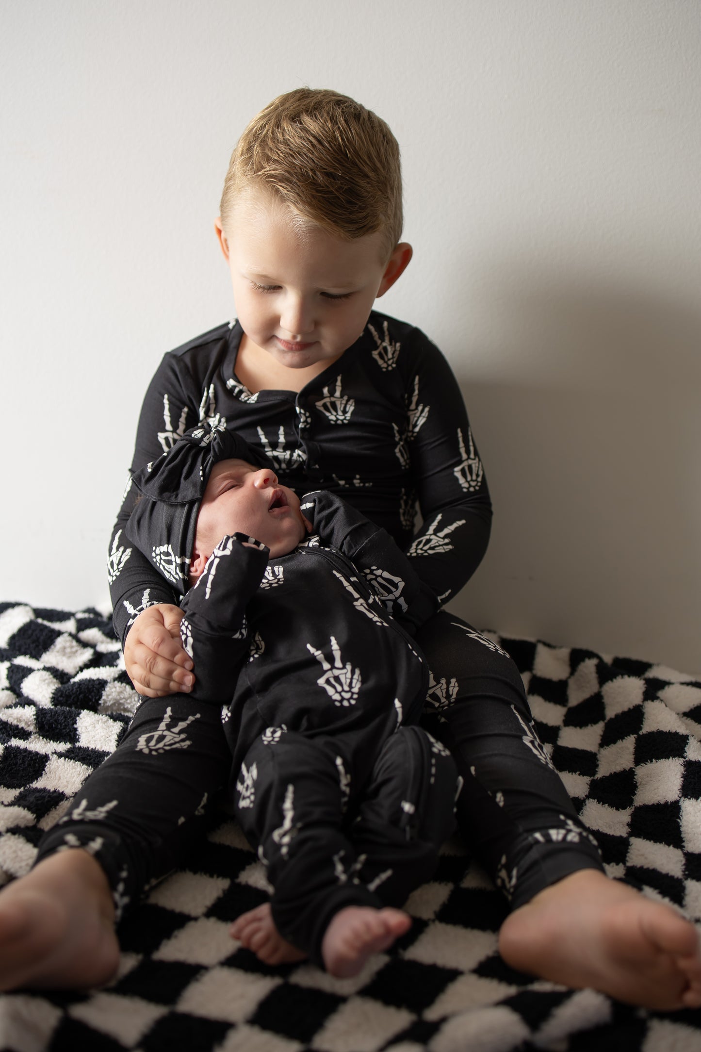 A young child in forever french baby’s Bamboo Two Piece Pajamas | Midnight Skelly sits on a checkered blanket, gently holding a baby dressed in matching bamboo pajamas and hat. Both are looking down as soft lighting highlights the comfort and breathability of their bamboo clothing.
