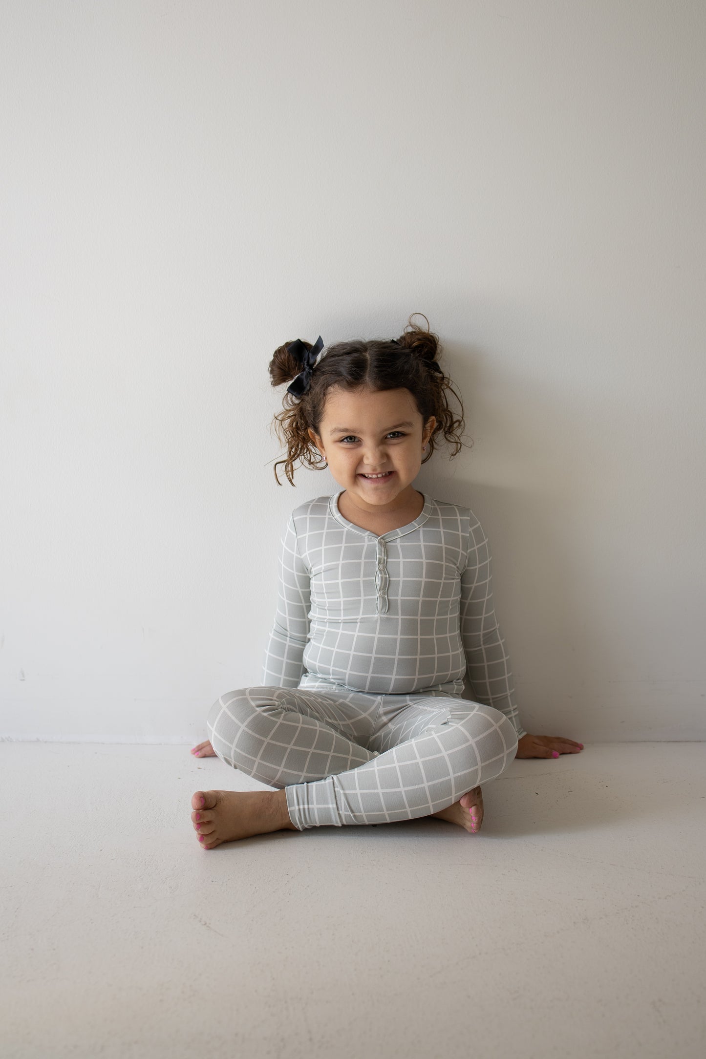 A young child with curly hair in pigtails sits on the floor against a plain wall, smiling and wearing the Bamboo Two Piece Pajamas | Foggy Grid by forever french baby. These light gray pajamas feature a white grid pattern and are made from hypo-allergenic bamboo fabric, making them perfect for sensitive skin while providing both comfort and style.