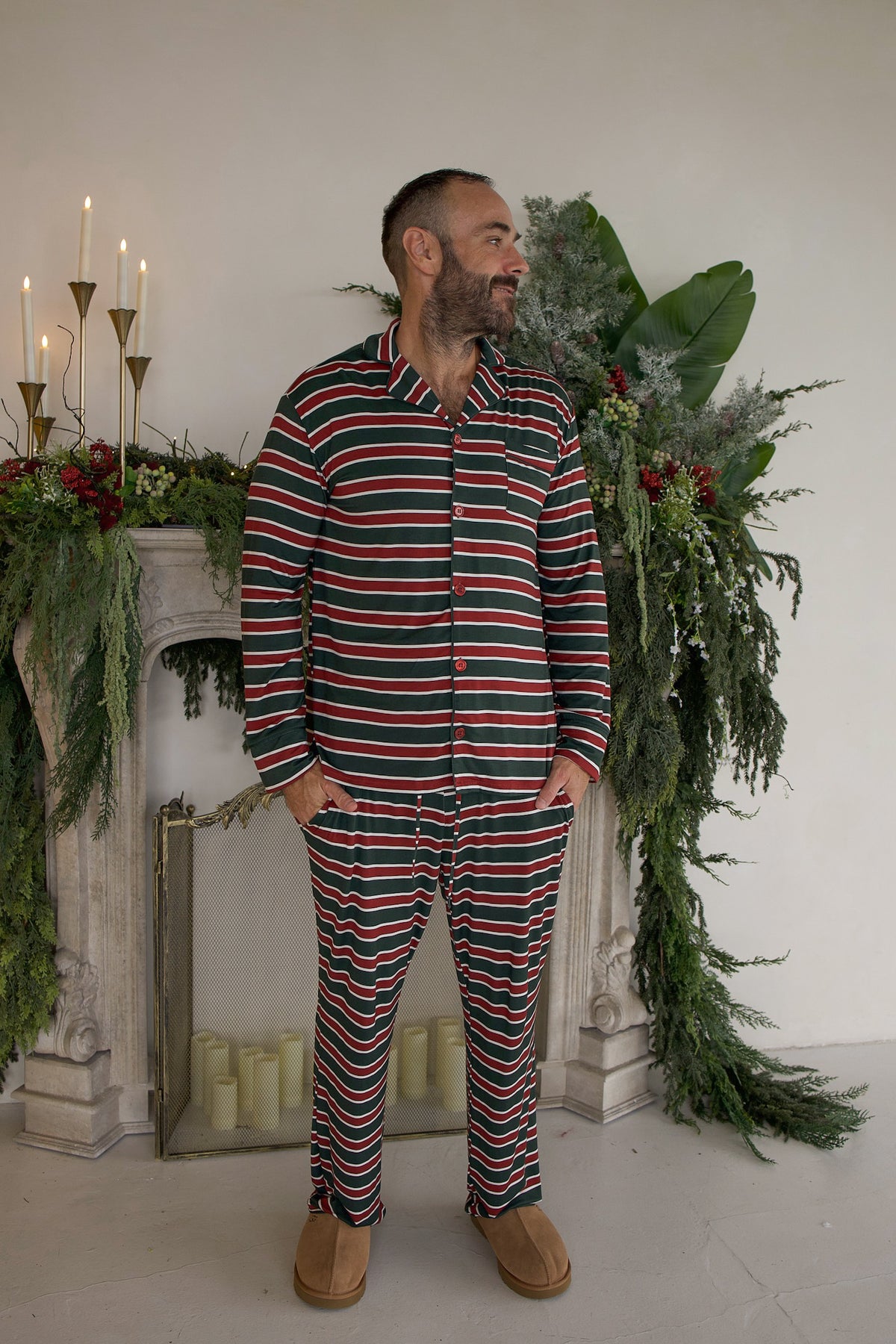 A man wearing the Men’s Bamboo Button Up Lounge Set | Holiday Stripe, consisting of a long-sleeved button-up shirt and matching lounge pants featuring horizontal red, green, and white stripes.