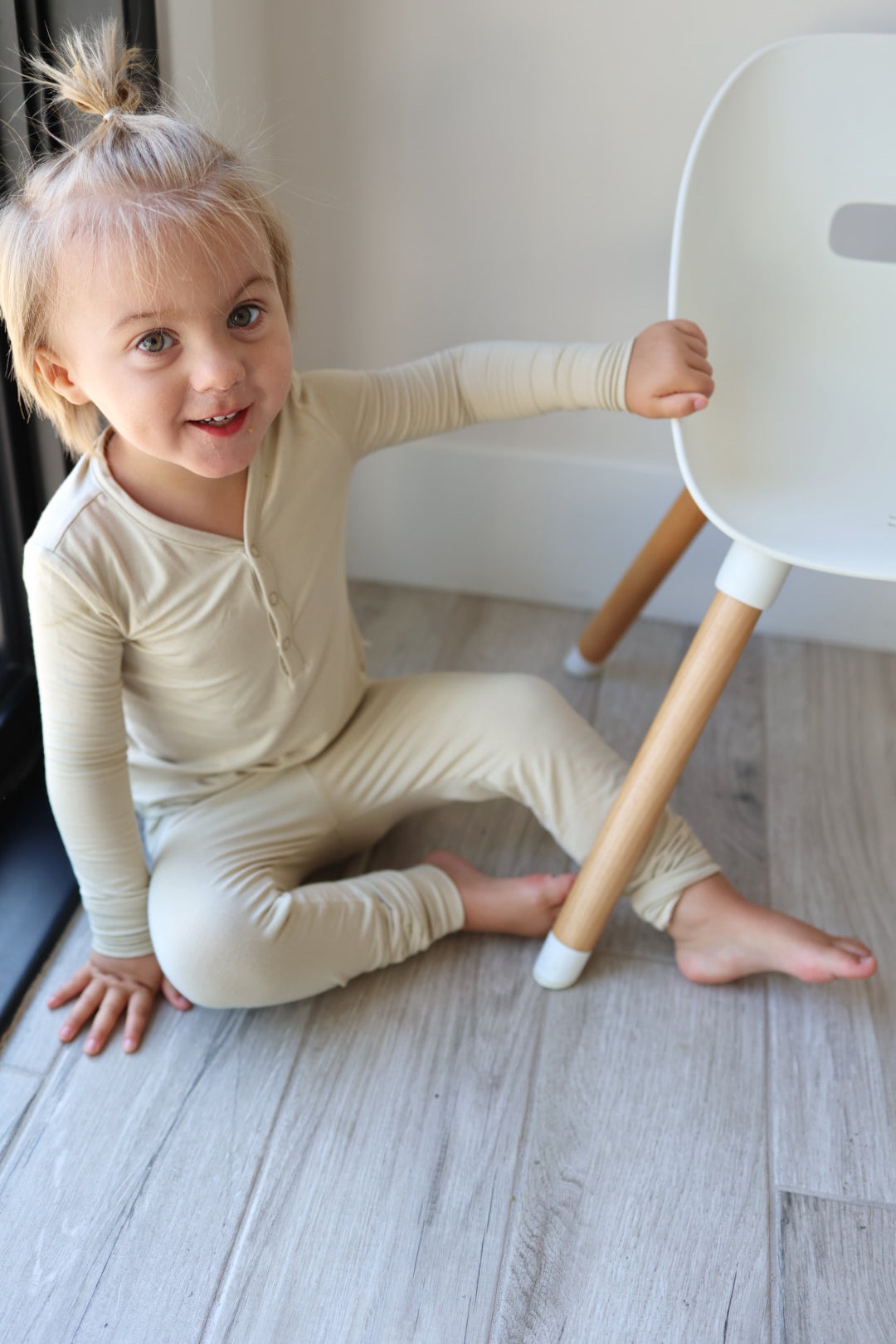 A young child with light-colored hair in a topknot sits on a wooden floor, wearing the Bamboo Two Piece Pajamas in Limestone from forever french baby. The breathable beige outfit compliments their bright smile as they hold onto the leg of a white chair beside them.