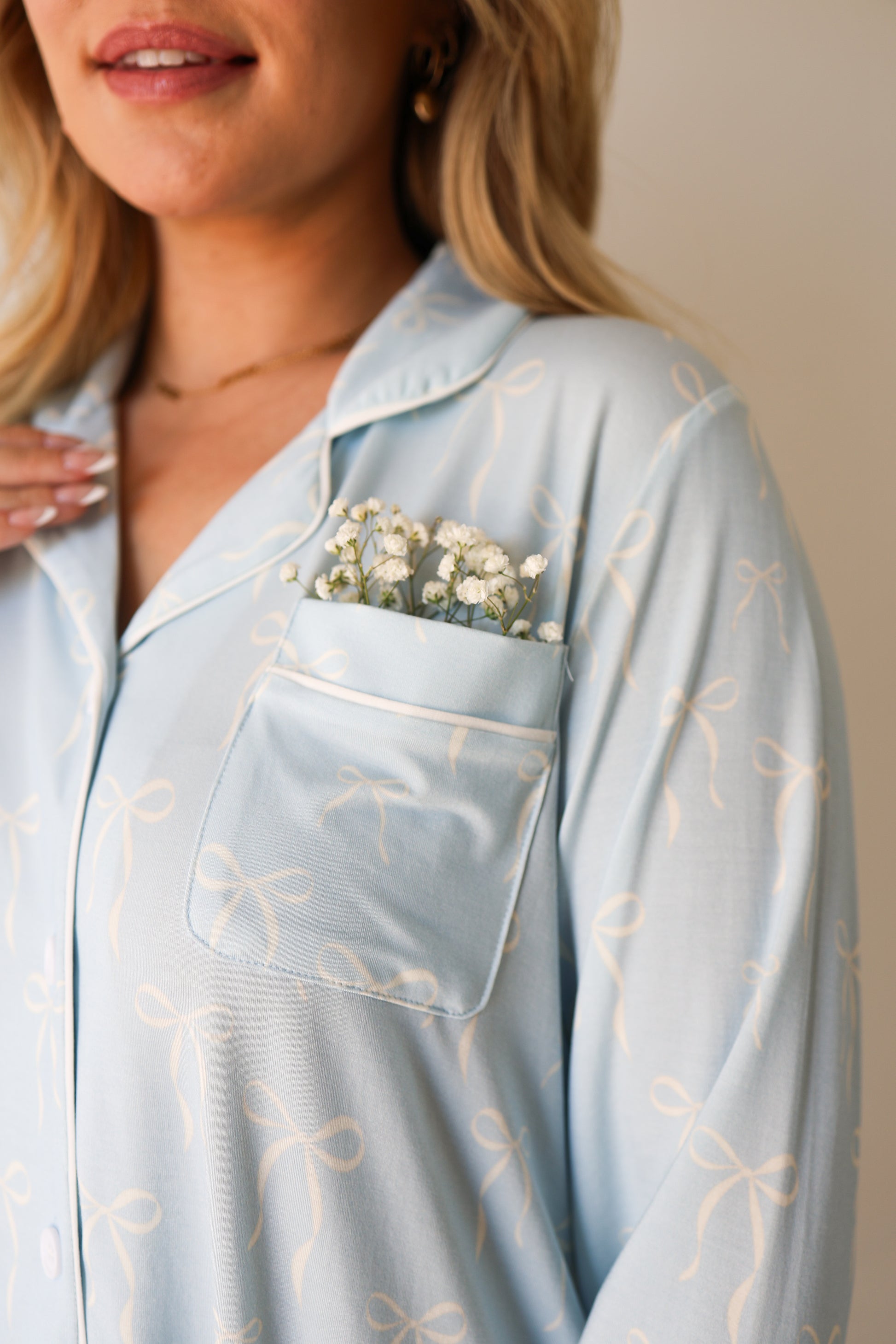 A woman wears the forever french baby Women's Bamboo Button Up Lounge Set in Blue Bows, featuring small white flowers tucked into the chest pocket. Only her lower face and upper torso are shown.