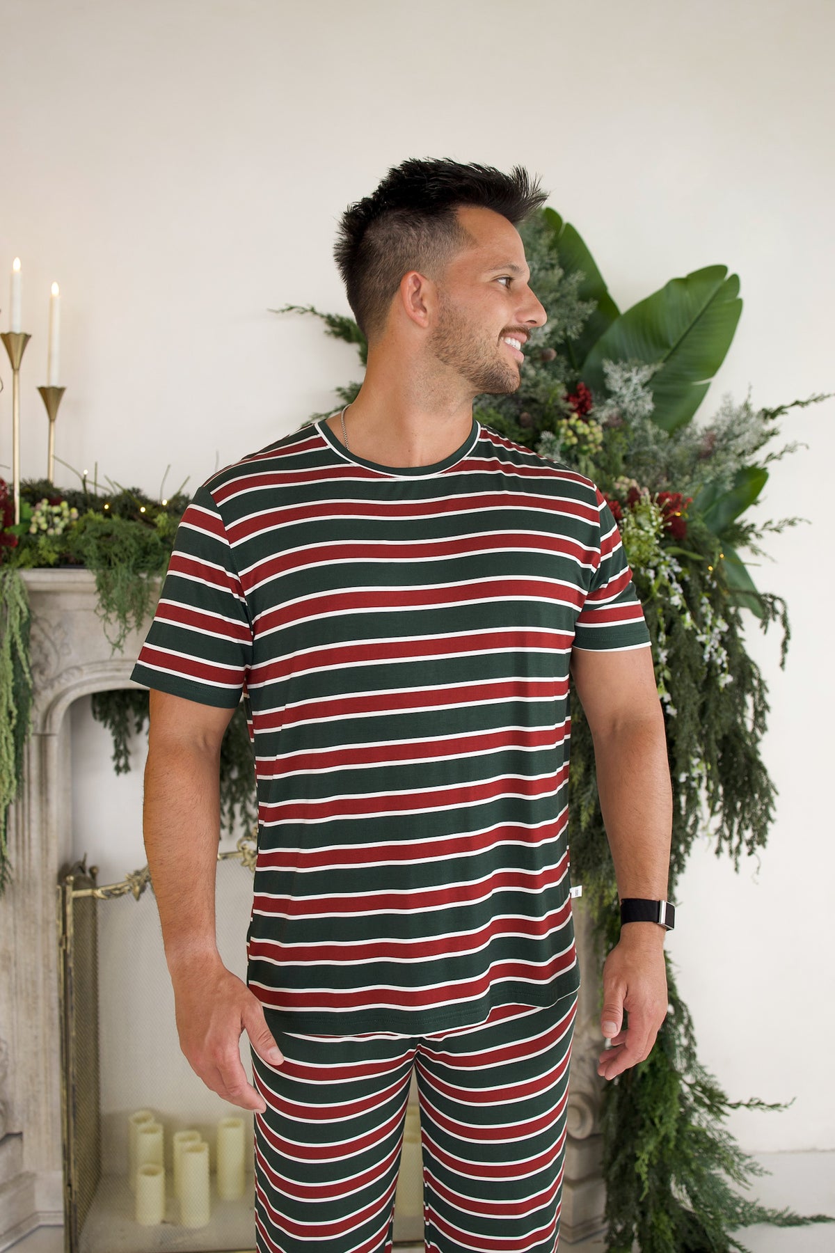 A man wears a Men’s Short Sleeve Pajamas | Holiday Stripe set, featuring a short-sleeve crew neck shirt and matching shorts with horizontal red, green, and white stripes.