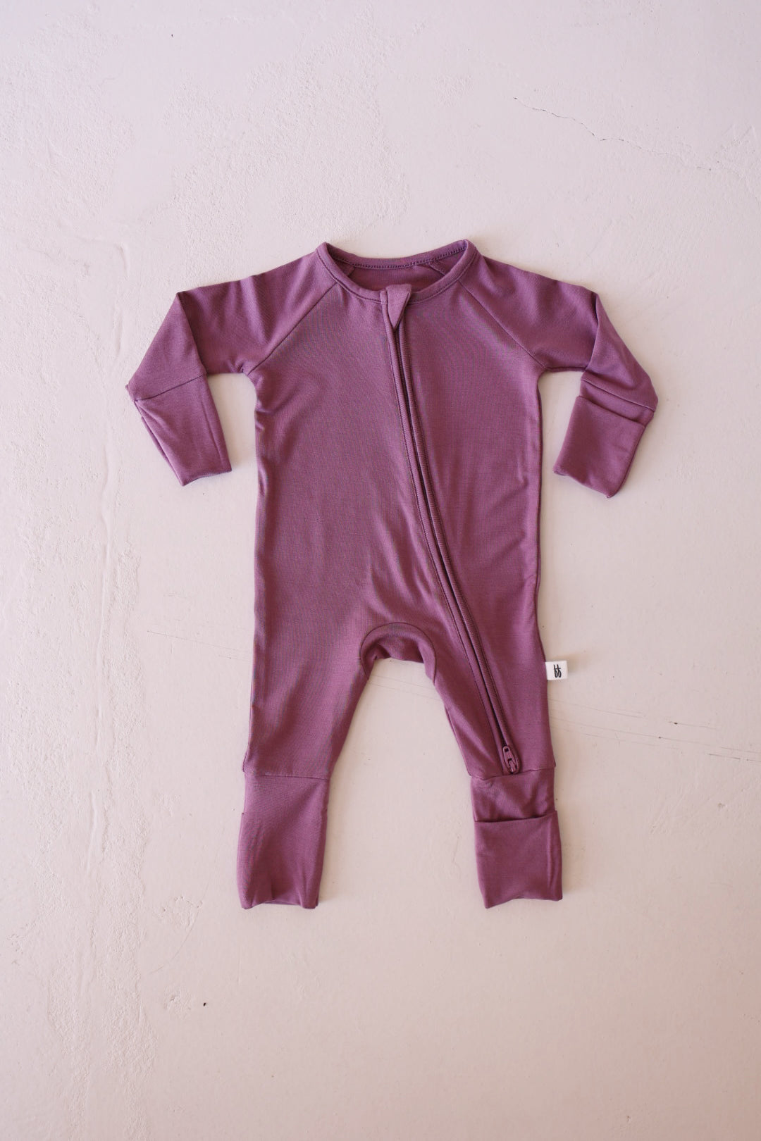 The Bamboo Zip Pajamas | Amethyst by forever french baby is a mauve-colored baby onesie with long sleeves and footed legs, crafted from breathable bamboo. It features a diagonal zipper running from the neck to the left leg and a small white tag on the left side seam. This hypo-allergenic onesie is laid flat on a light background.