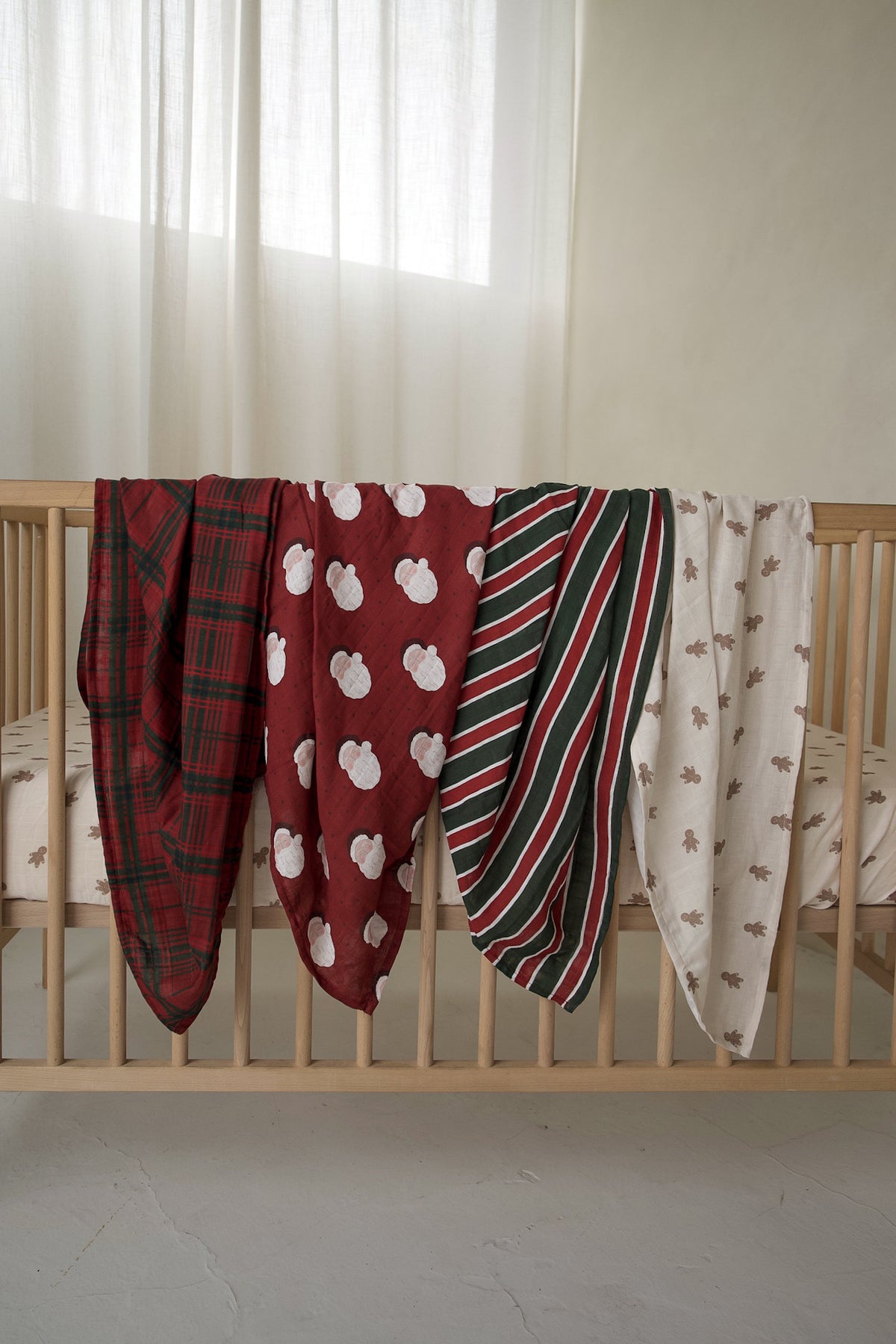 Four ultra-soft Muslin Swaddle | Christmas Plaid blankets with red, green, and white holiday patterns draped over a wooden crib.