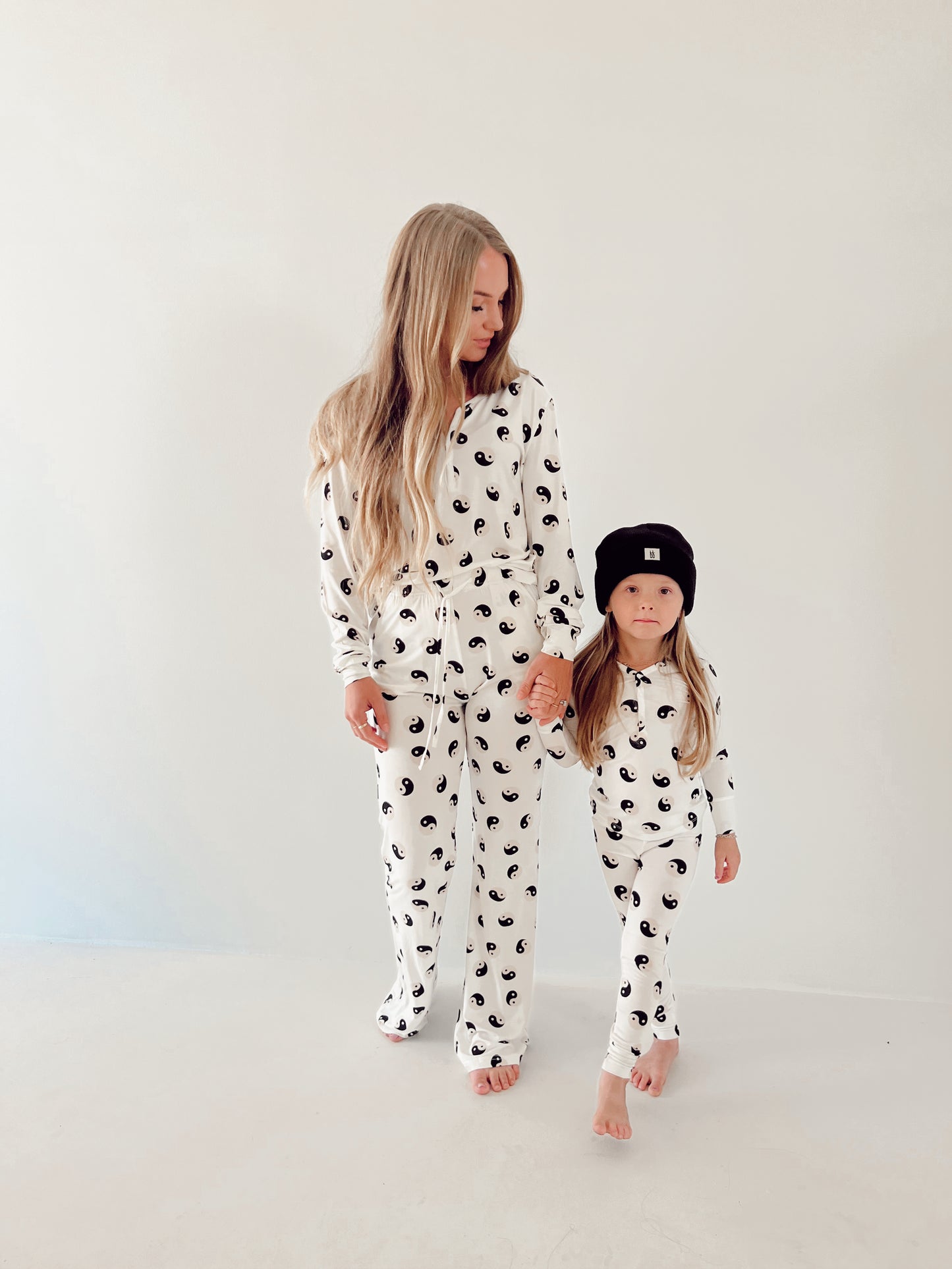 A woman with long blonde hair and a child holding hands stand against a plain white background, wearing matching "Women's Bamboo Pajamas | Black & White Yin Yang" by forever French baby. The child is also wearing a black beanie, and both are barefoot.