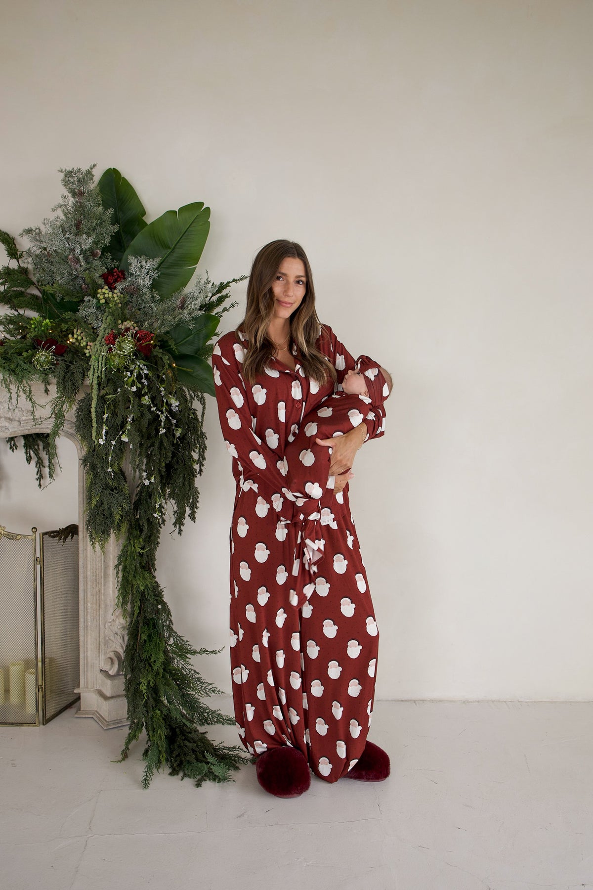A woman wears a maroon Women’s Bamboo Button Up Lounge Set | Vintage Santa with a repeating white Santa Claus print, including wide leg lounge pants, while holding a newborn wrapped in a matching swaddle.