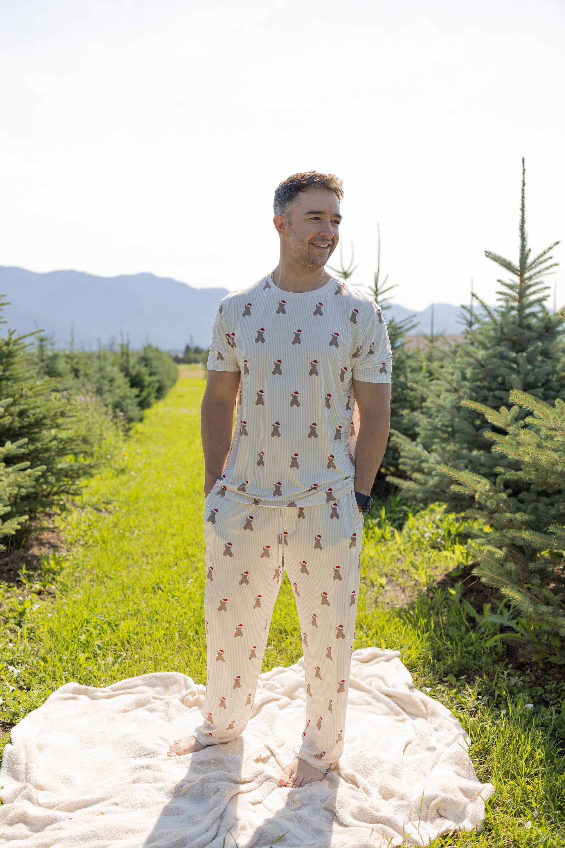 A man stands barefoot on a blanket in a grassy field between rows of evergreen trees, wearing the forever french baby Men's Short Sleeve Pajamas in the Santa Teddy print. Mountains are visible in the background under a bright sky.