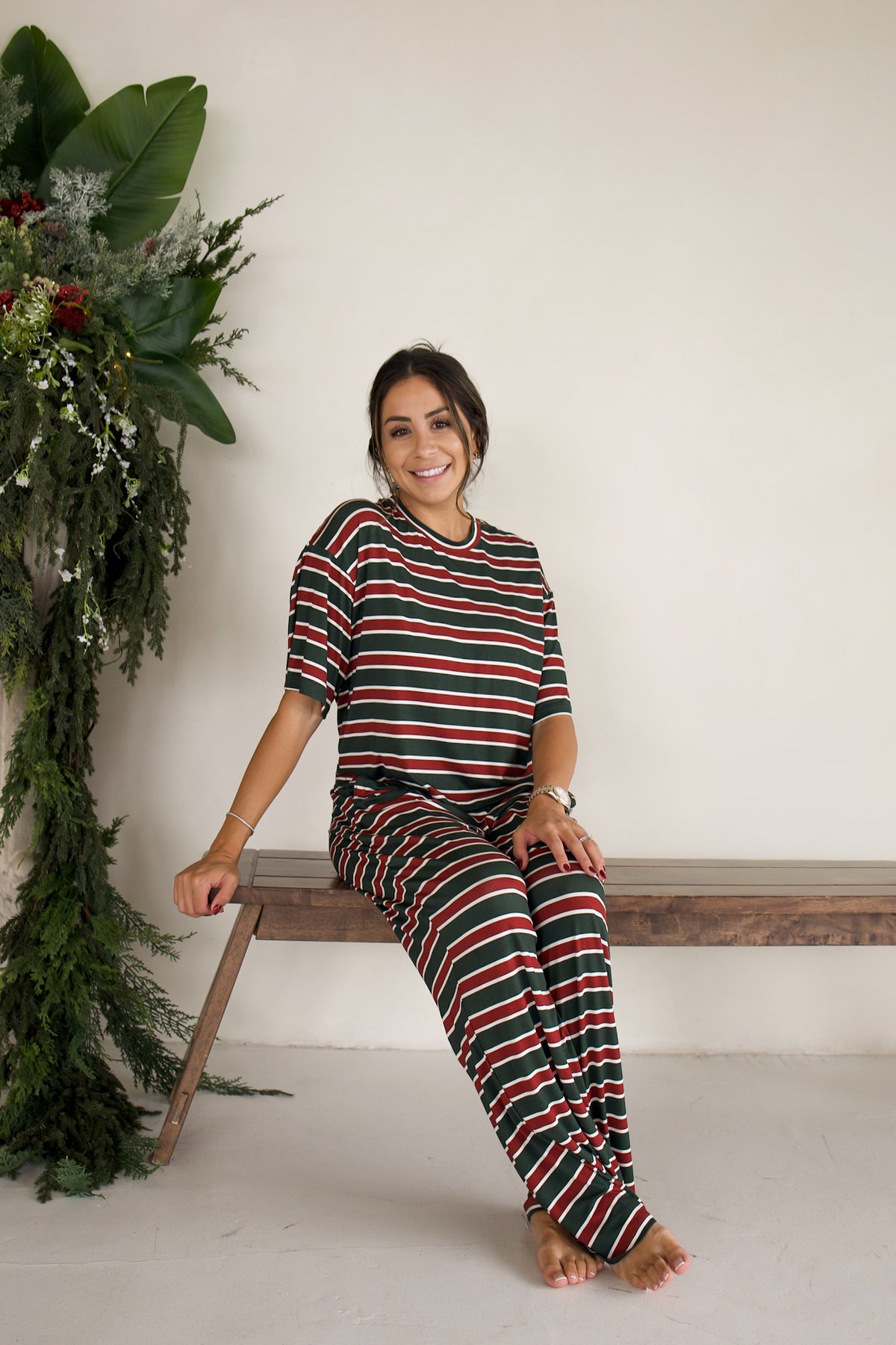 A woman sits on a wooden bench wearing a forever french baby Women’s Boxy Tee Lounge Set | Holiday Stripe, featuring a matching short-sleeved top and wide-leg pants in red, green, and white horizontal stripes.