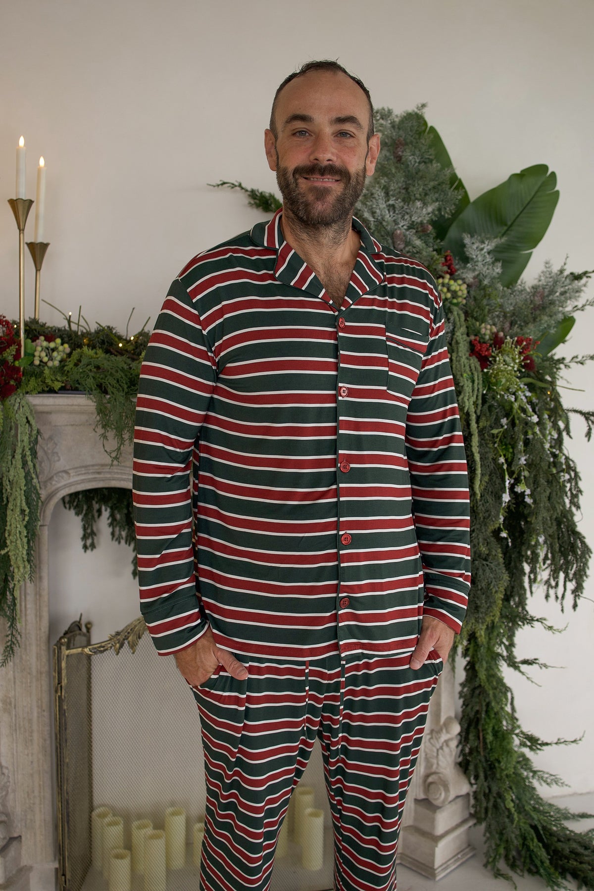 A man wearing the forever french baby Men’s Bamboo Button Up Lounge Set | Holiday Stripe, featuring bamboo lounge pants and a matching long-sleeved button-up shirt in a red, green, and white horizontal stripe pattern.