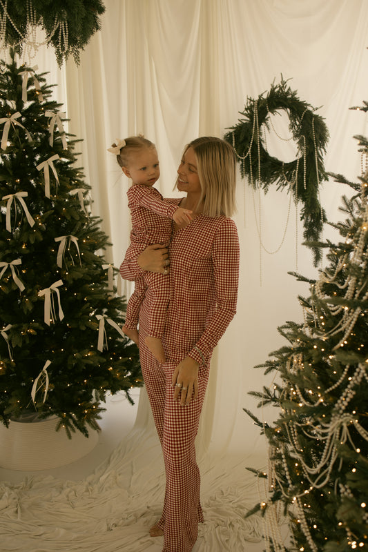 A woman in forever french baby's Women's Bamboo Pajamas | Red Gingham holds a young girl in matching sleepwear by decorated Christmas trees, a wreath, white drapes, and festive lights.
