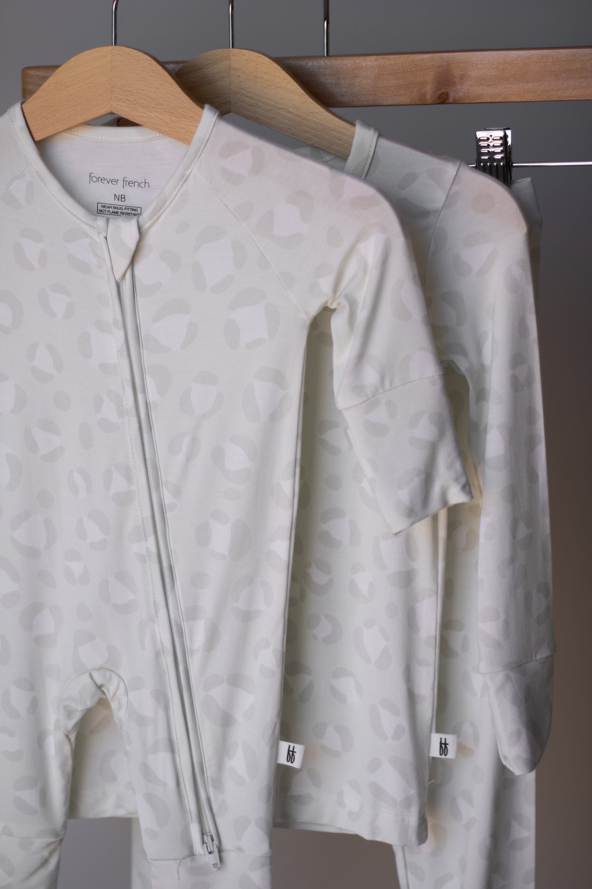 Two pale, patterned Bamboo Zip Pajamas | Meow by forever french baby hang on wooden hangers. Made from breathable fabric, these onesies feature subtle abstract prints and front zippers for comfort and style.
