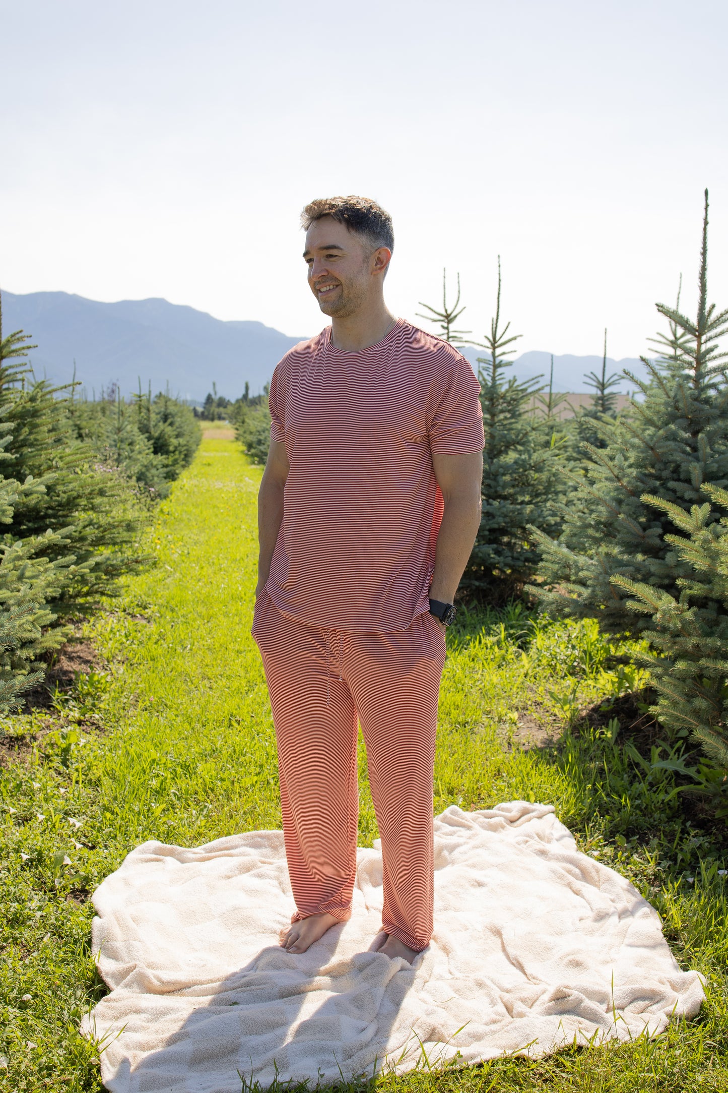 A man in the Men's Short Sleeve Pajamas in Peppermint Stripe Ribbed by forever french baby stands on a beige blanket between rows of pine trees in a sunny outdoor setting, with mountains in the background.